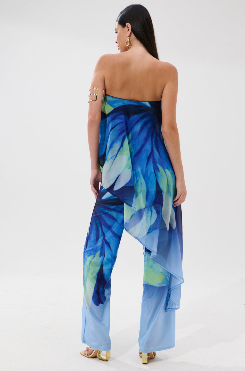 A woman faces away in the ISLAND QUEEN JUMPSUIT, featuring a strapless, flowy silhouette and bold abstract florals. Styled with gold hoop earrings, gold heels, and long dark hair—ideal for a chic getaway look.