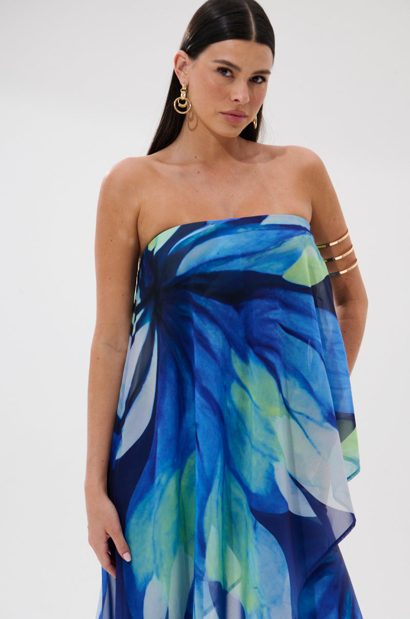 A woman with long dark hair wears the ISLAND QUEEN JUMPSUIT, a strapless, flowy piece with a bold blue and green floral print. She pairs it with gold hoop earrings and a gold arm cuff, standing against a plain white background.
