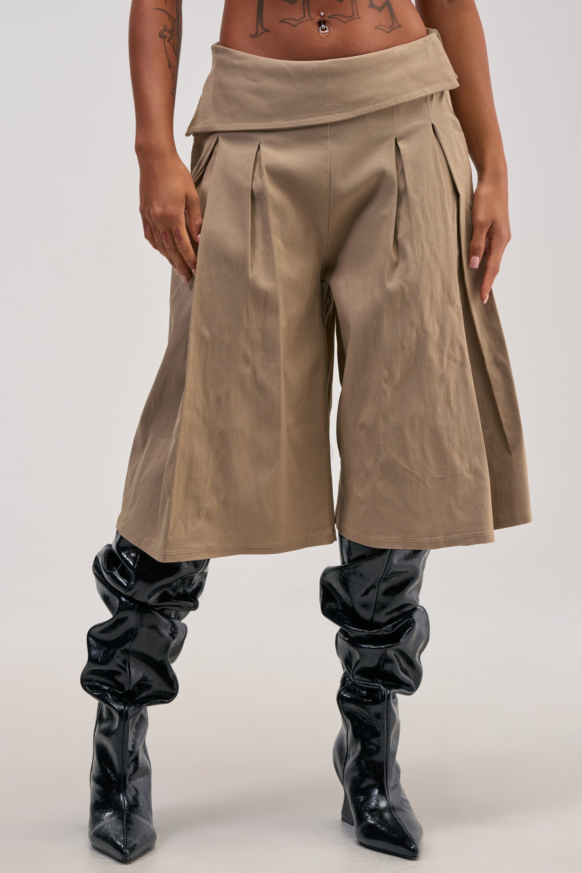 Wearing the DOTTIE BERMUDA SHORT—tan, pleated, and wide-leg with a folded waistband—the person pairs them with shiny black slouchy knee-high boots. With hands resting on their hips, tattoos along their torso and arms highlight the high-rise fit of the shorts.