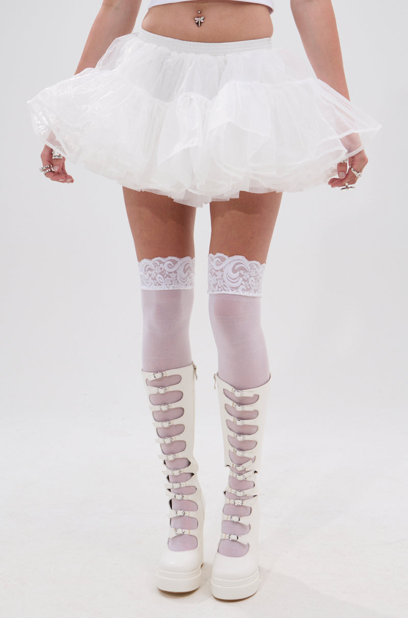 Wearing the JENNY TULLE MINI SKIRT, white lace thigh-high stockings, and high-rise knee-high boots, this person stands against a plain background. Their visible midriff and belly button piercing complete a stylish festival look. 1