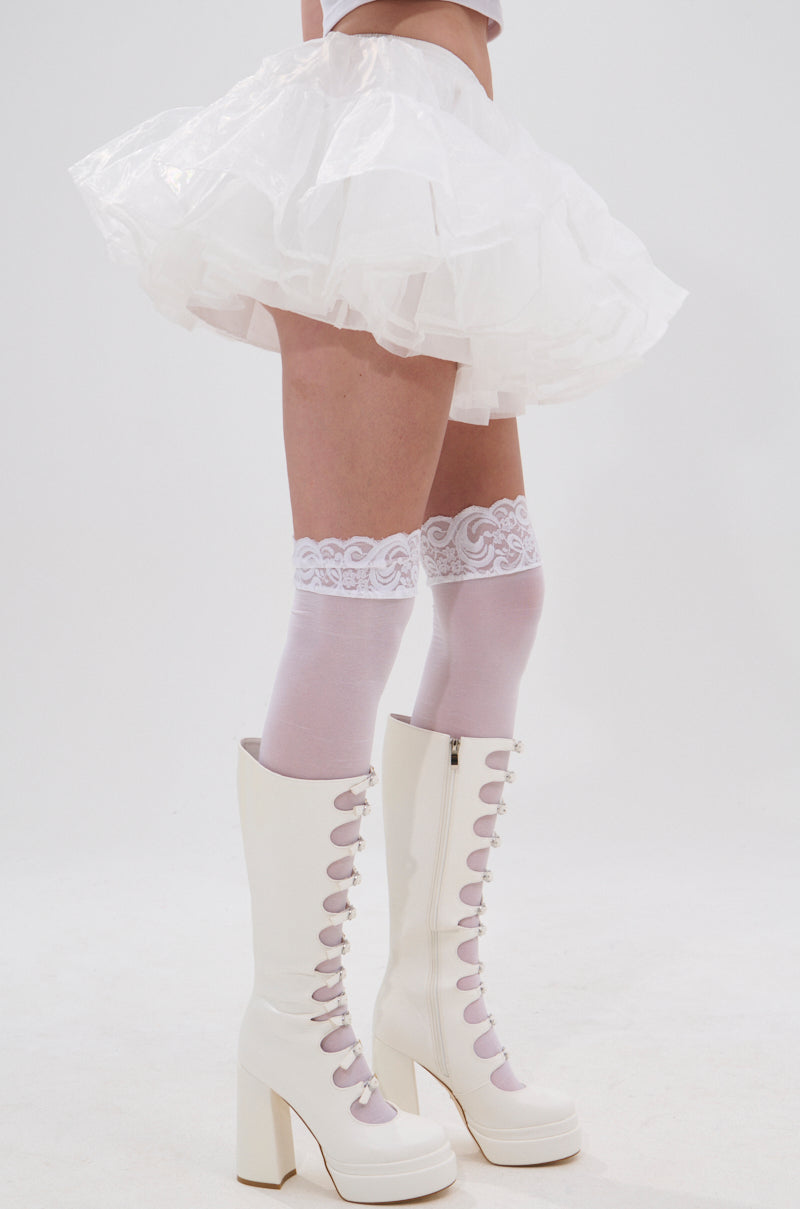Wearing the JENNY TULLE MINI SKIRT, knee-high lavender socks with lace trim, and white high-heeled lace-up boots, a person stands against a plain white background in this festival-ready look. 2