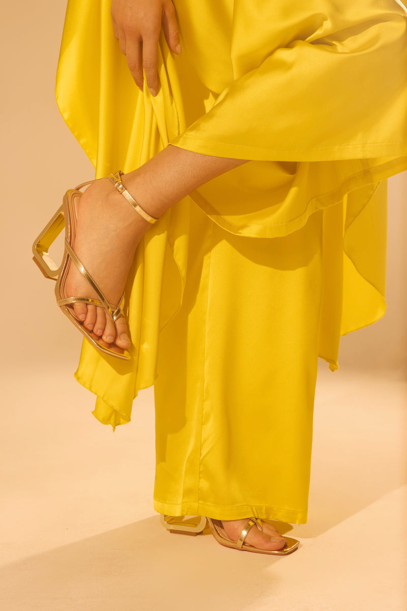 A person in a flowing yellow dress lifts the hem to reveal AKIRA BRANWEN GOLD WEDGE SANDALS with a Comfort Cushion insole, set against a soft beige background.