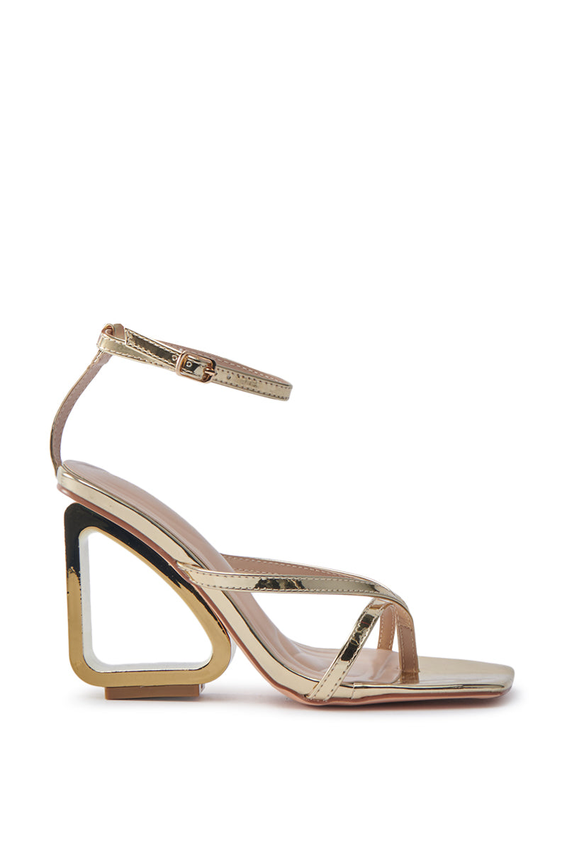The AKIRA BRANWEN GOLD WEDGE SANDAL is a gold high-heeled shoe with a unique sculpted geometric cut-out heel, featuring thin straps, an ankle strap with a buckle, and an open toe for an elegant and modern look.