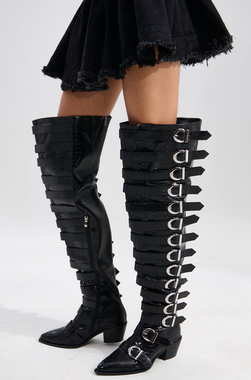 Someone models the AZALEA WANG BRAULIA BLACK CROC WESTERN BOOT, featuring black croc-embossed faux leather, chunky cowboy heels, pointed toes, and buckled straps, paired with a black frayed-hem skirt against a plain light background.