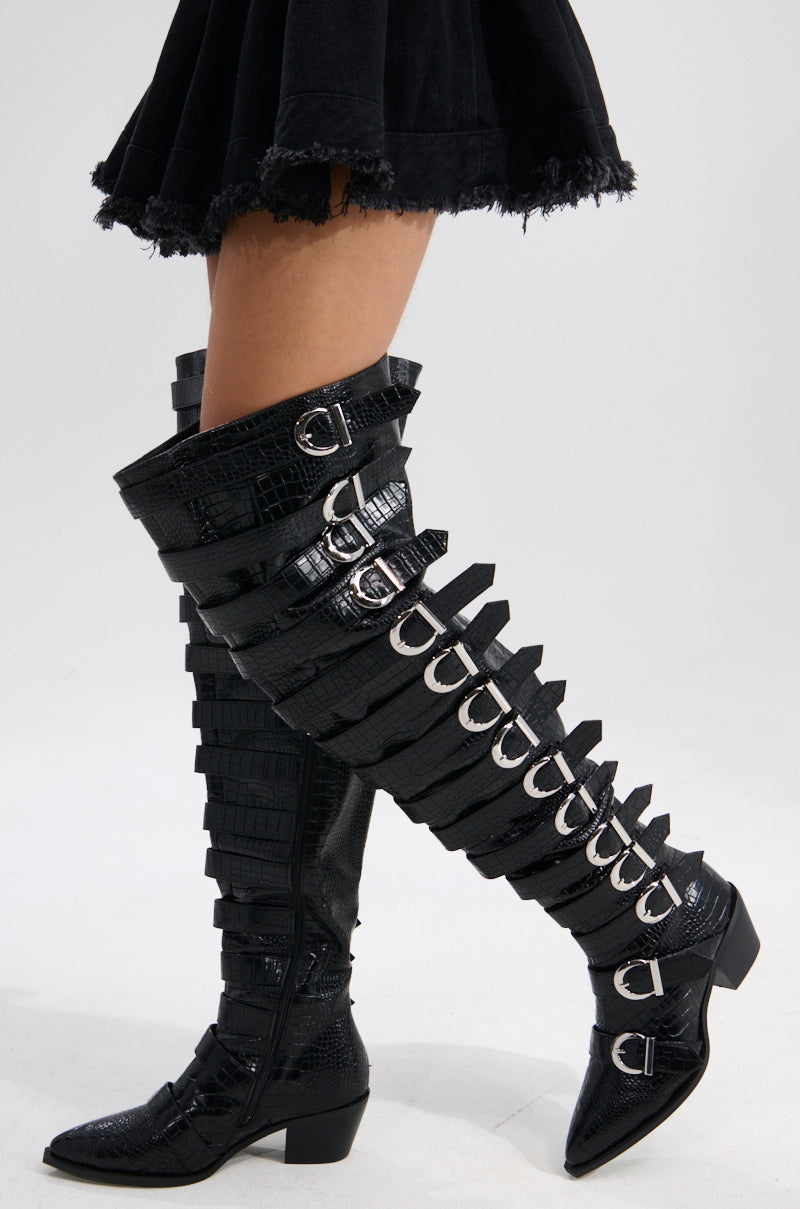 A person wears the AZALEA WANG BRAULIA BLACK CROC WESTERN BOOT—thigh-high, black croc, with multiple silver-buckled straps, a chunky heel, and a pointed toe—paired with a black skirt.