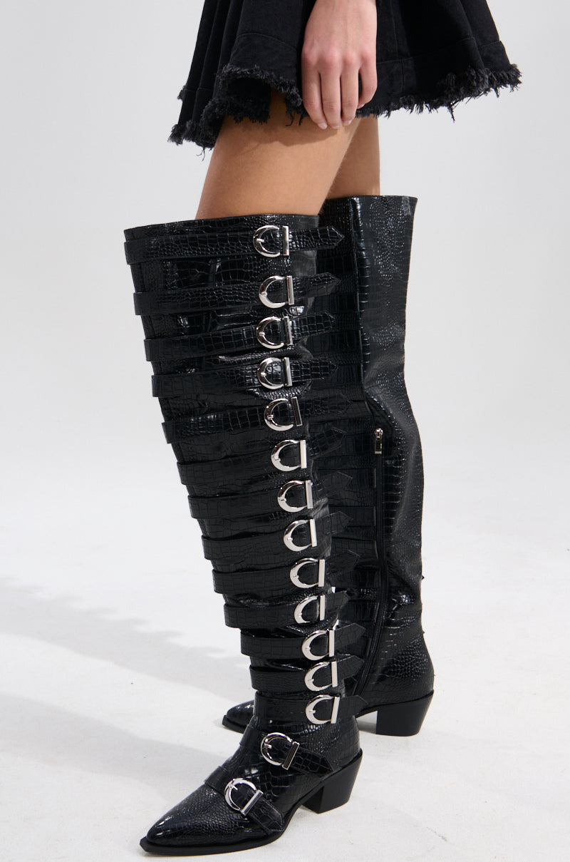 A person in a black skirt wears AZALEA WANG BRAULIA BLACK CROC WESTERN BOOTS with chunky heels and multiple silver buckles, standing against a plain light gray background.