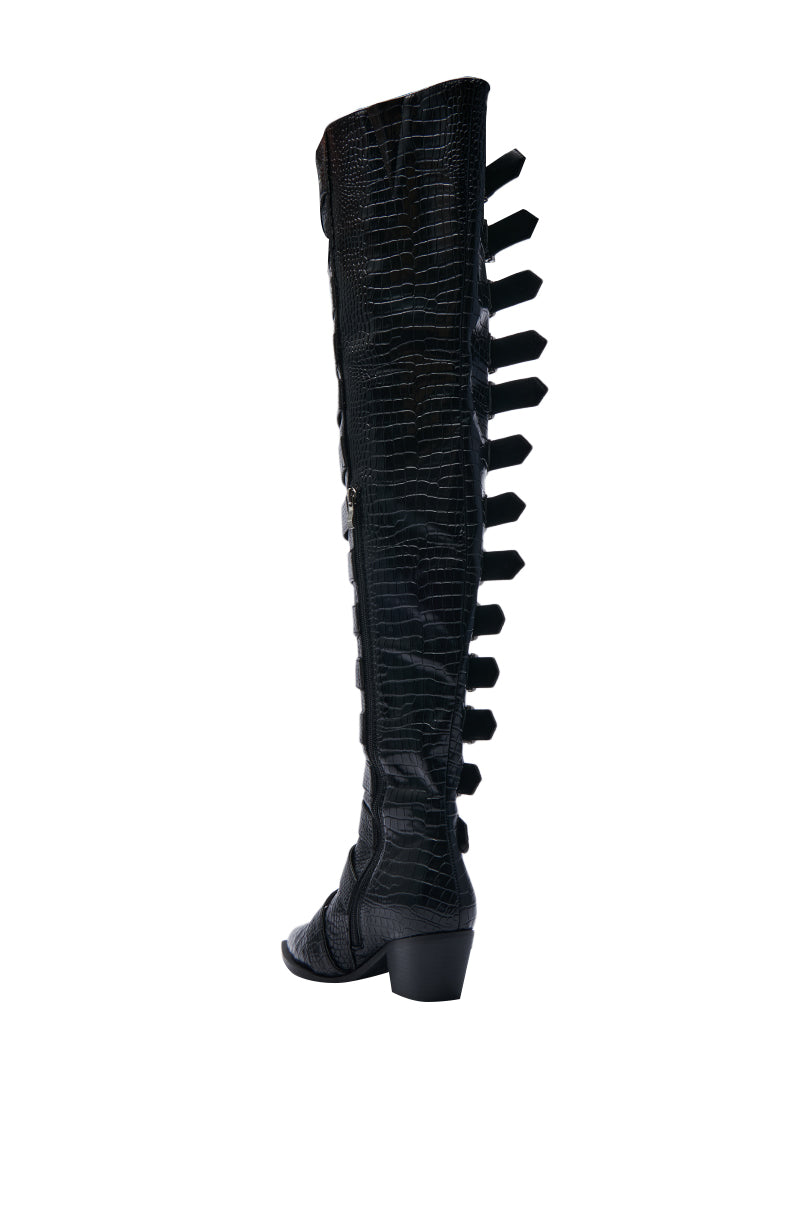 A single knee-high boot in black, with a faux crocodile skin texture and a convenient side zipper. It showcases black rectangular cutouts running down its length, complemented by a low chunky heel. This sophisticated AZALEA WANG BRAULIA BLACK CROC WESTERN BOOT is displayed against a white background.