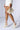 A person is walking in a chic, white short outfit with a pearl-adorned handbag and matching AZALEA WANG BREAN Embellished Pumps featuring an abstract inverted wedge heel. The person's short sheer socks add an additional texture to the elegant ensemble.