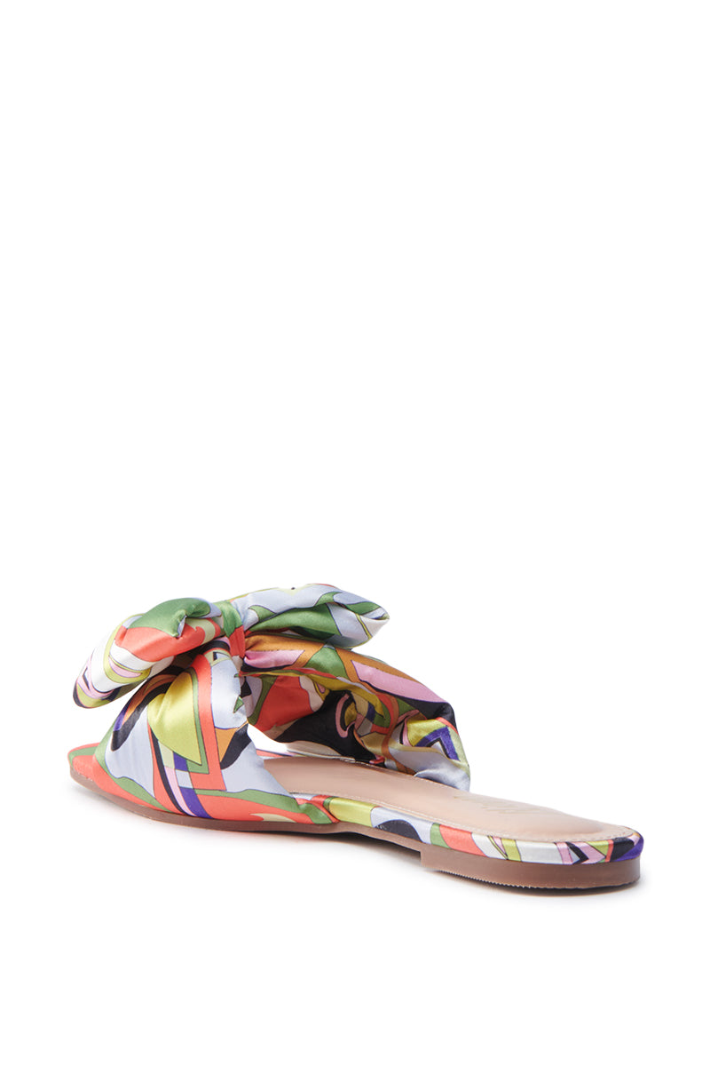 The BREEZY SCARF FLAT SANDAL IN CHARTREUSE features a vibrant abstract pattern in orange, green, blue, and yellow, adorned with a large decorative bow on top. It includes a tan insole and flat sole for comfort.