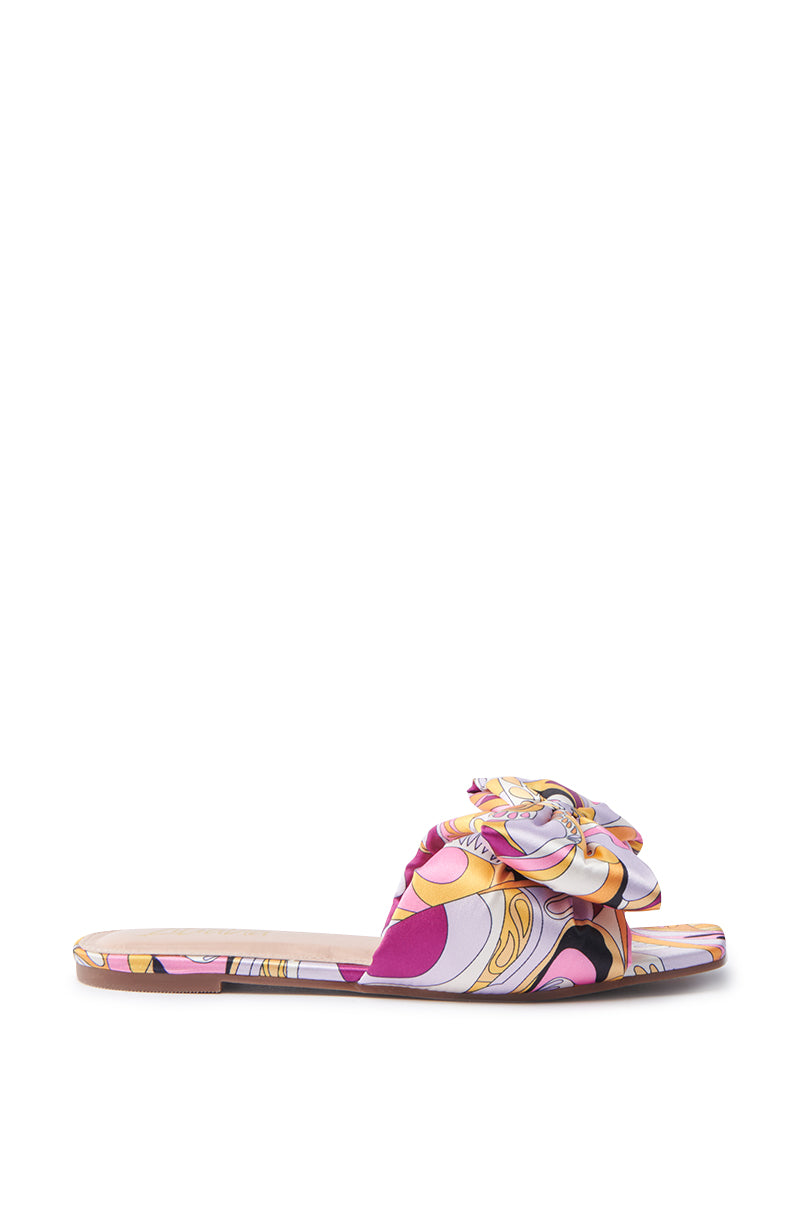 The BREEZY SCARF FLAT SANDAL IN FUCHSIA is a vibrant slide sandal, featuring a flat sole and open toe. Its satin upper displays a swirling pattern in pink, orange, yellow, and purple with a decorative fabric flower.