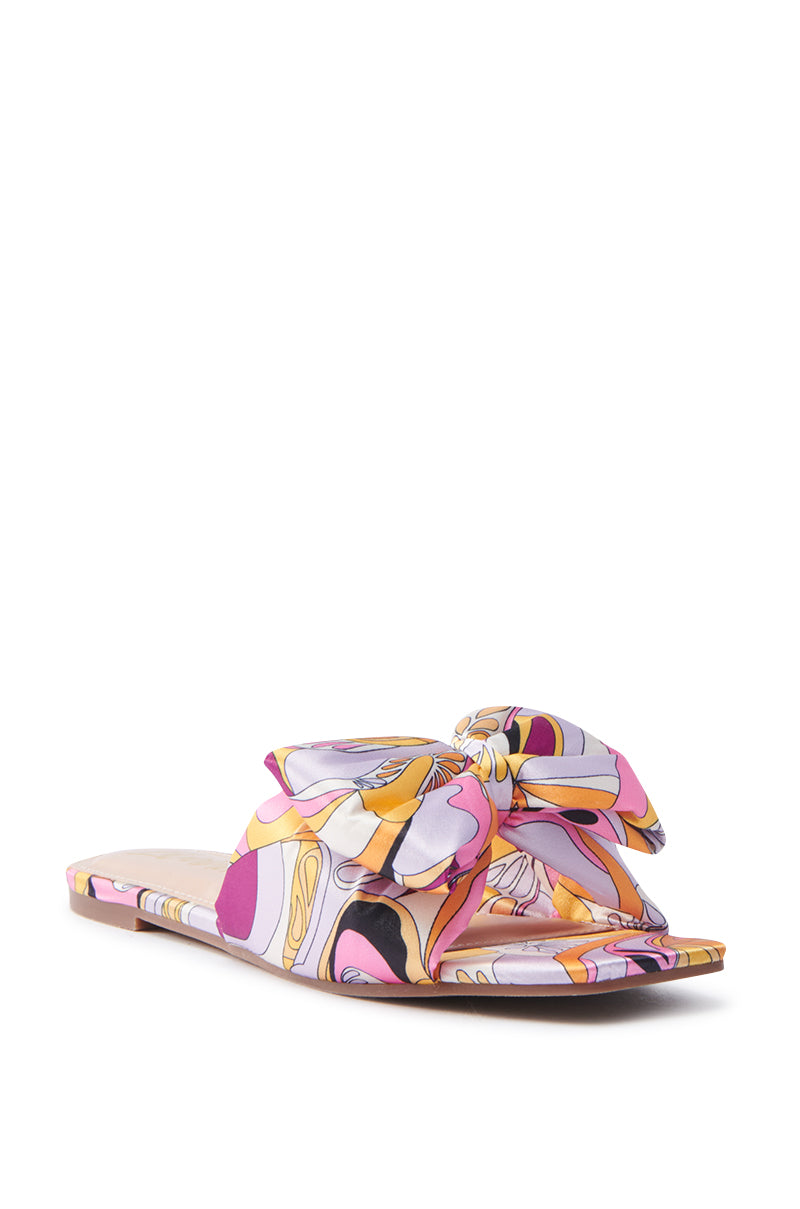 The BREEZY SCARF FLAT SANDAL IN FUCHSIA features a pink, yellow, and white abstract floral print on satin with a large knotted bow on the strap. Its square toe design and flat sole provide a casual yet stylish appeal.