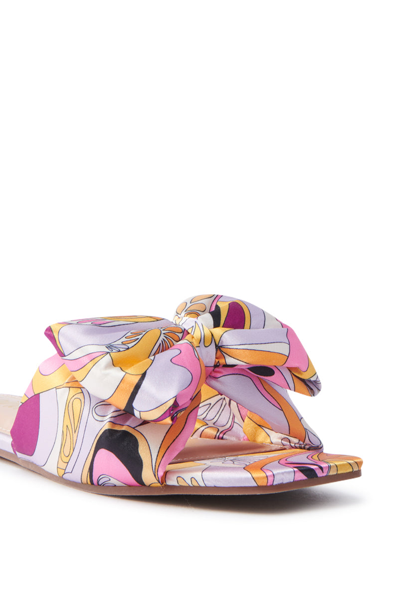 A close-up of the BREEZY SCARF FLAT SANDAL IN FUCHSIA showcases a square toe slide with a patterned bow. Swirling shapes in pink, purple, and yellow adorn the sleek satin upper, elegantly standing out against a plain white background.