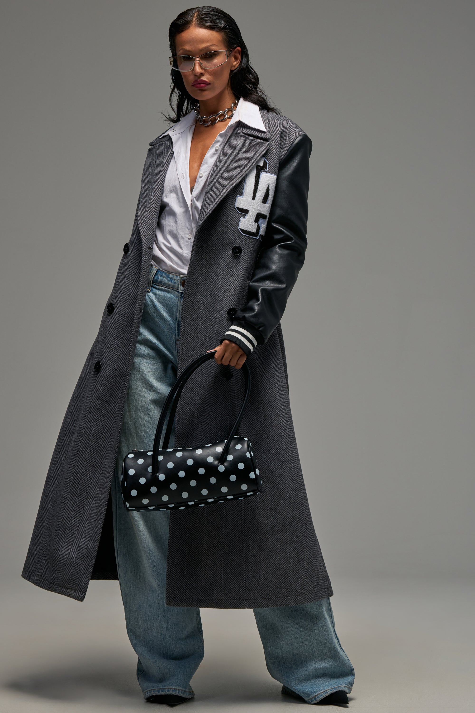 A fashionable person wearing the HIDDEN HILLS HERRINGBONE VARSITY TRENCH—a chic grey herringbone coat with black leather sleeves—paired with a white shirt, blue wide-leg jeans, a black and white polka dot bag, red glasses, and a chunky necklace.