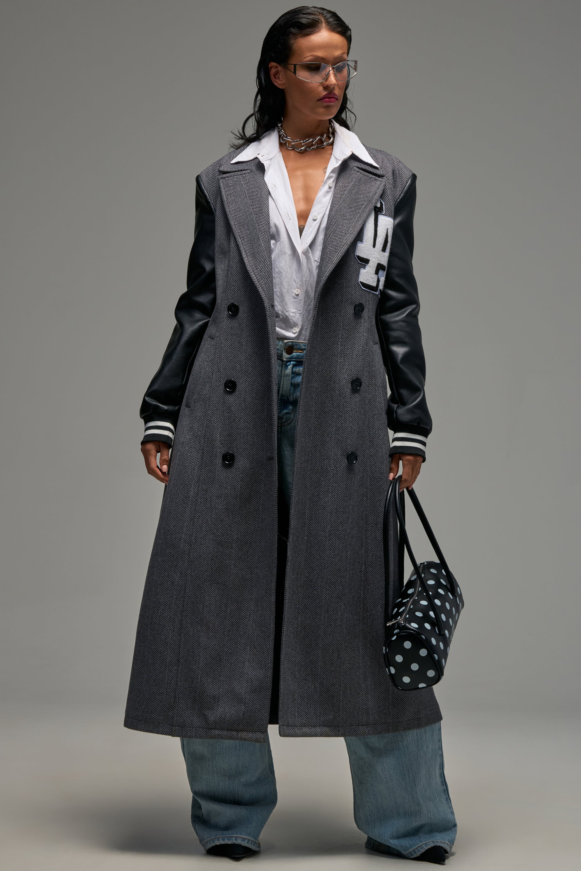 Wearing the HIDDEN HILLS HERRINGBONE VARSITY TRENCH in stylish grey with black leather sleeves, paired with a white shirt, wide-leg jeans, and glasses, this person holds a black and white polka-dot bag while standing against a plain background.