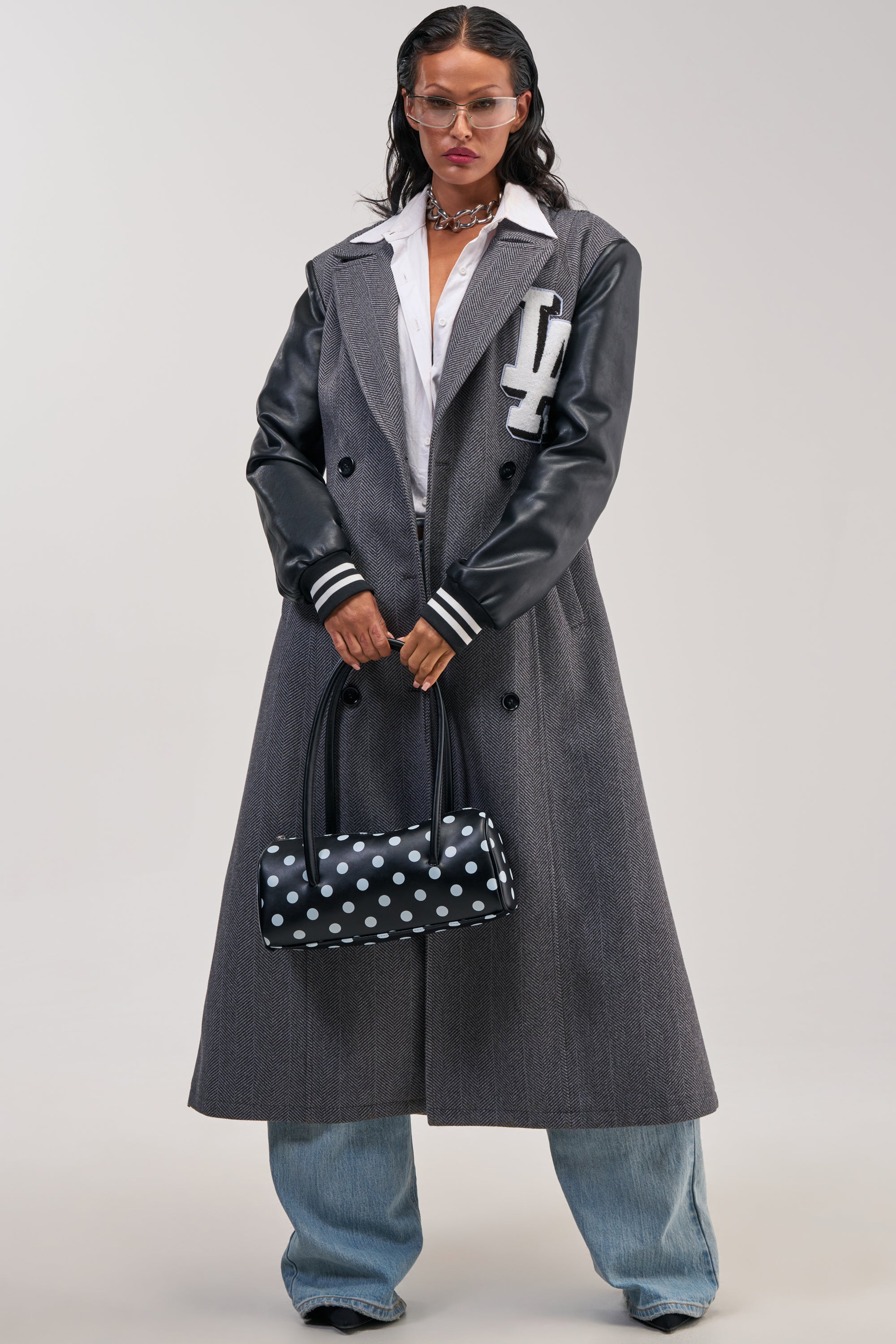A person wearing the HIDDEN HILLS HERRINGBONE VARSITY TRENCH, styled with blue jeans, a white shirt, black leather sleeves, pink-tinted glasses, and holding a black polka dot handbag stands against a plain background.