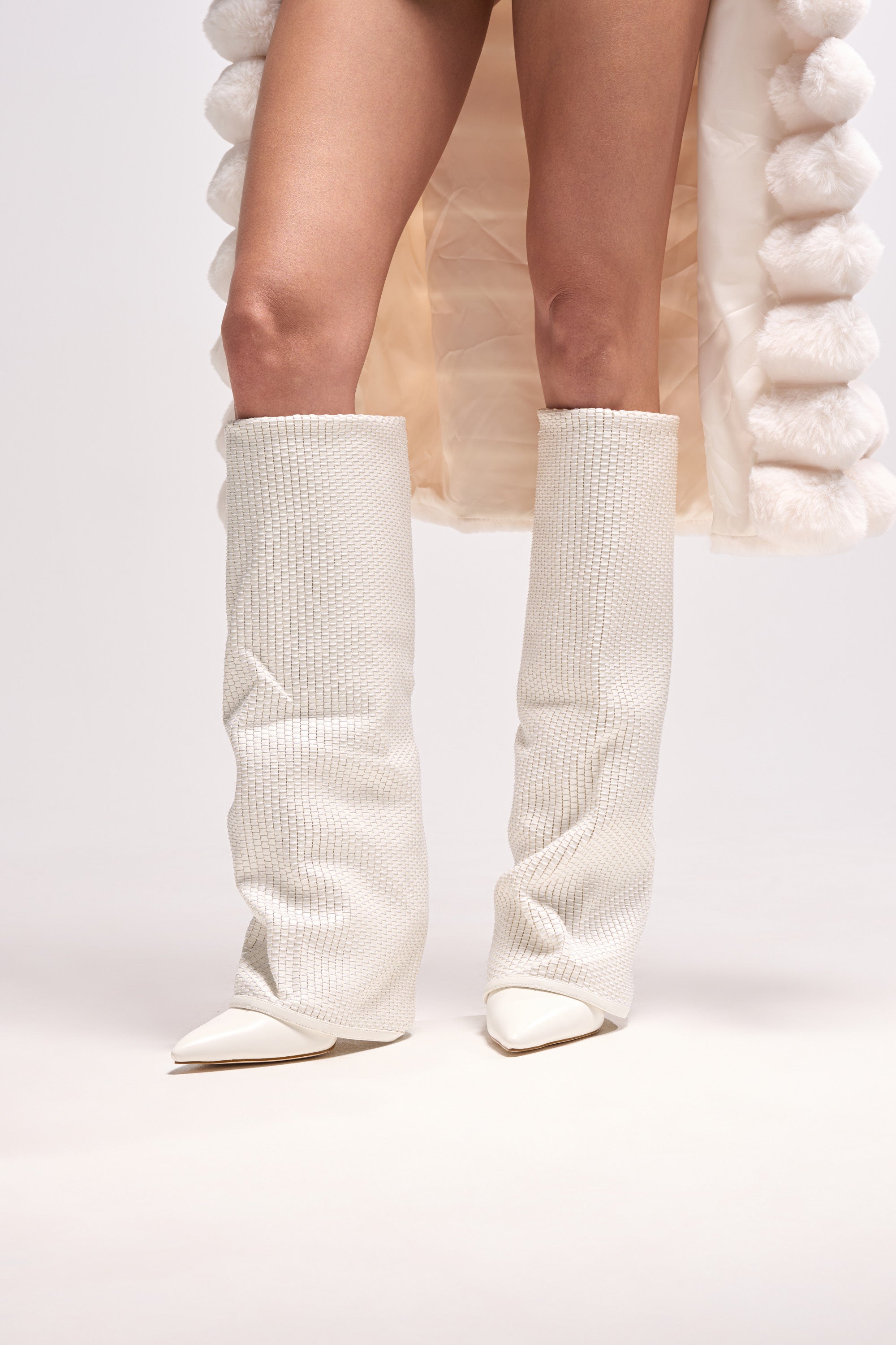 A person stands against a white background, showing only their legs and the lower part of a coat, wearing the AZALEA WANG BRETANGE WOVEN PU FOLD OVER BOOT IN WHITE. 1