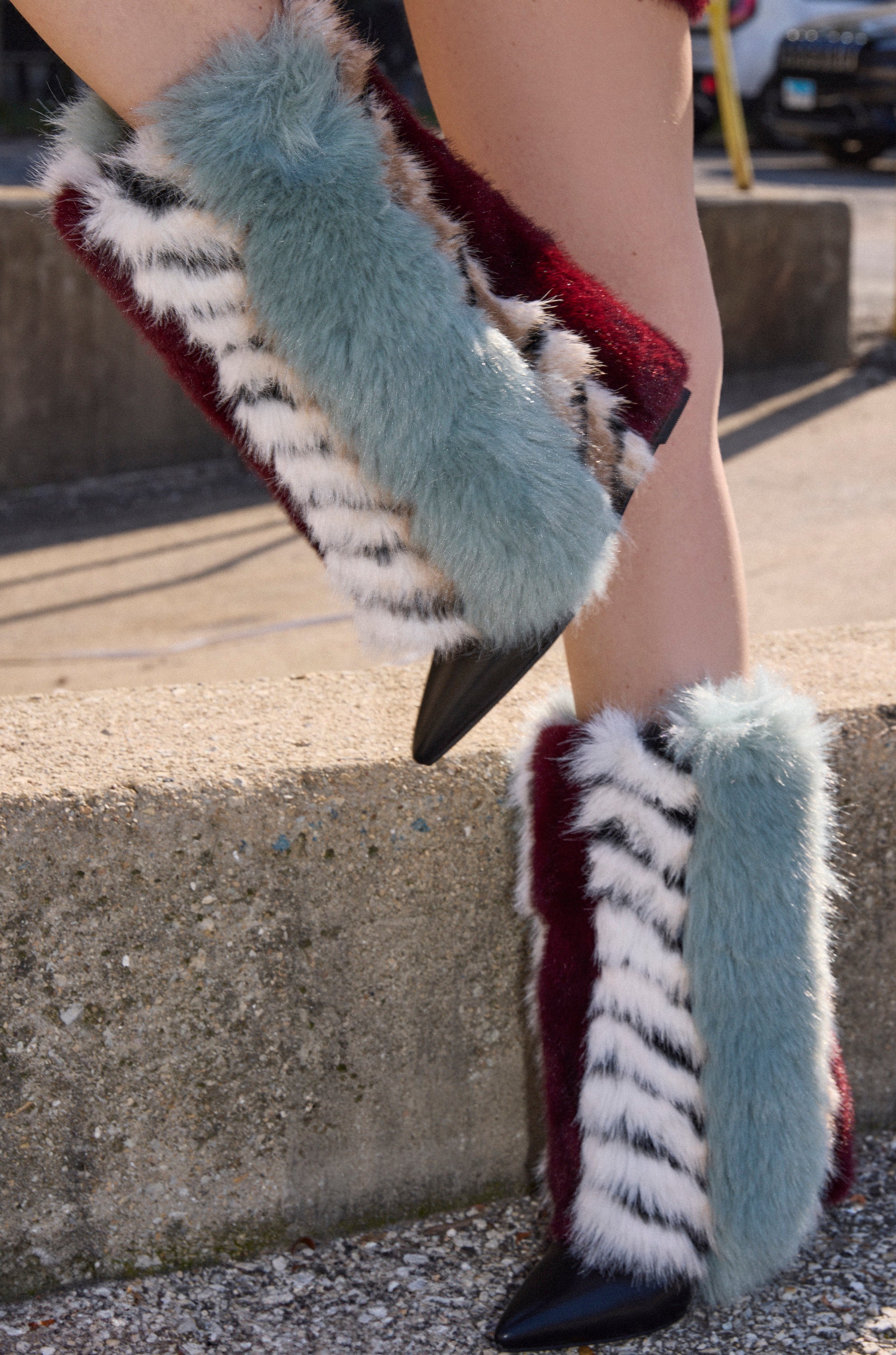 A close-up of two legs wearing the AZALEA WANG BRIASH BLUE MULTI FUR HIDDEN WEDGE BOOTIE, featuring multicolored fur stripes and pointed black toes, set outdoors against a concrete background.