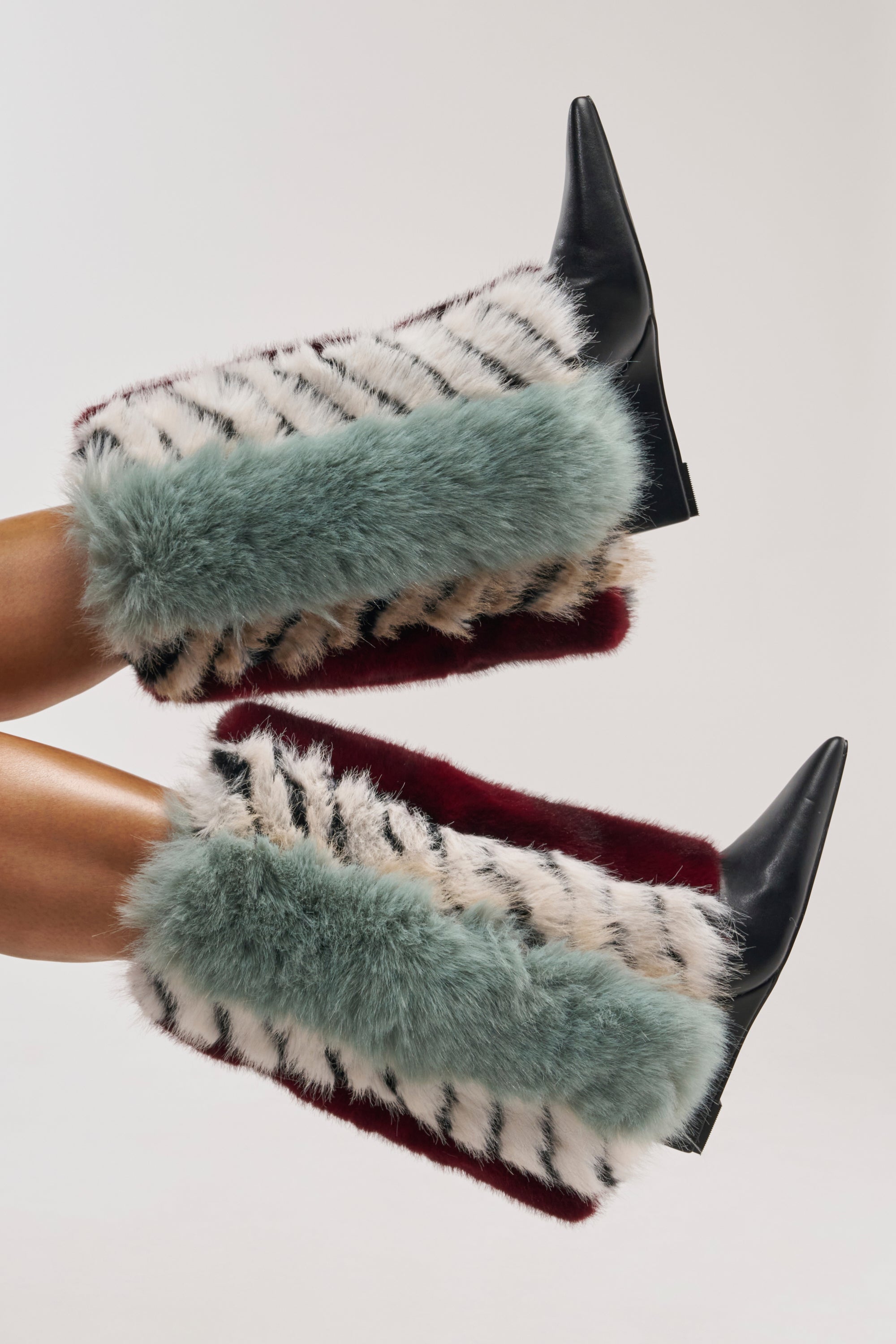 Two legs wearing the AZALEA WANG BRIASH BLUE MULTI FUR HIDDEN WEDGE BOOTIE—knee-high boots with a pointed toe, thick faux fur in teal, burgundy, and white accented by black stripes—stand against a plain light background. The boots showcase bold streetwear styling for a textured and vibrant look.