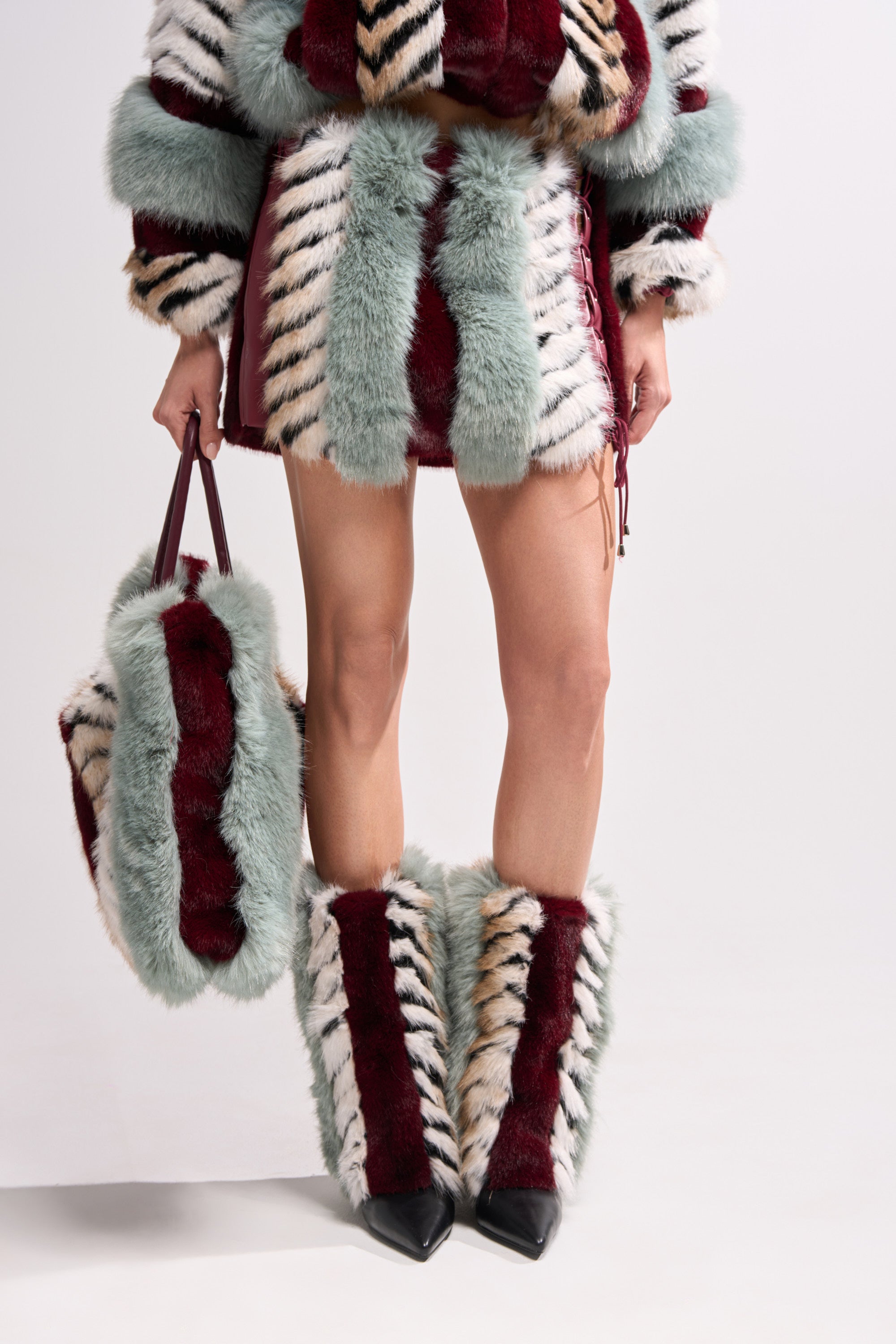 A person wears a striped faux fur mini skirt and tote, paired with the AZALEA WANG BRIASH BLUE MULTI FUR HIDDEN WEDGE BOOTIE in burgundy, mint green, black, and cream. The furry, fashion-forward boots are eye-catching; only the lower body is visible.