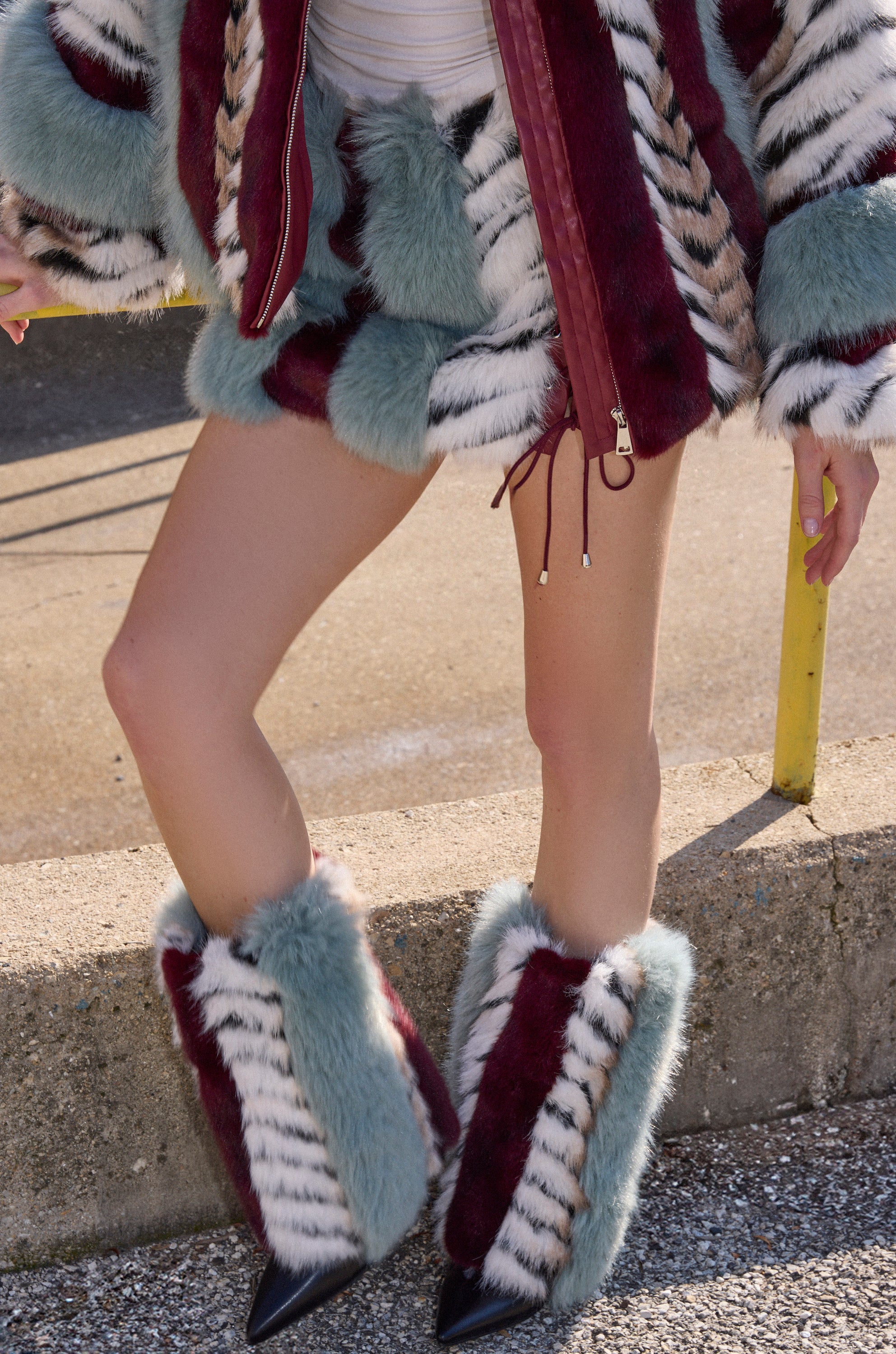 Only the legs and lower torso are visible of a person outdoors on concrete near a yellow railing, wearing AZALEA WANG BRIASH BLUE MULTI FUR HIDDEN WEDGE BOOTIE as part of a colorful faux fur look with blue, burgundy, and white striped boots.