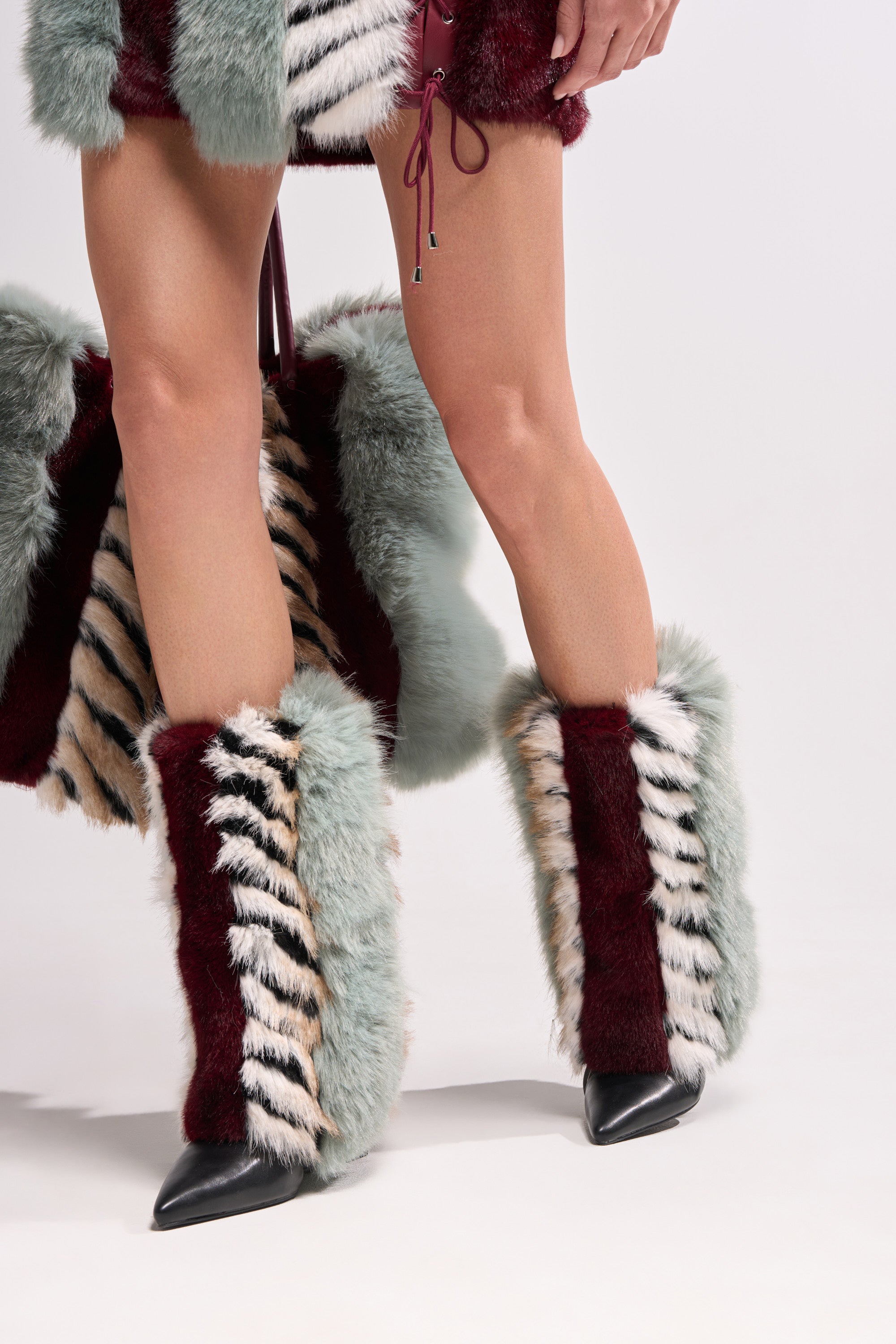 Wearing the AZALEA WANG BRIASH BLUE MULTI FUR HIDDEN WEDGE BOOTIE and a striped skirt in burgundy, mint green, cream, and black, a person stands against a plain light background with only their lower body and part of an arm visible.