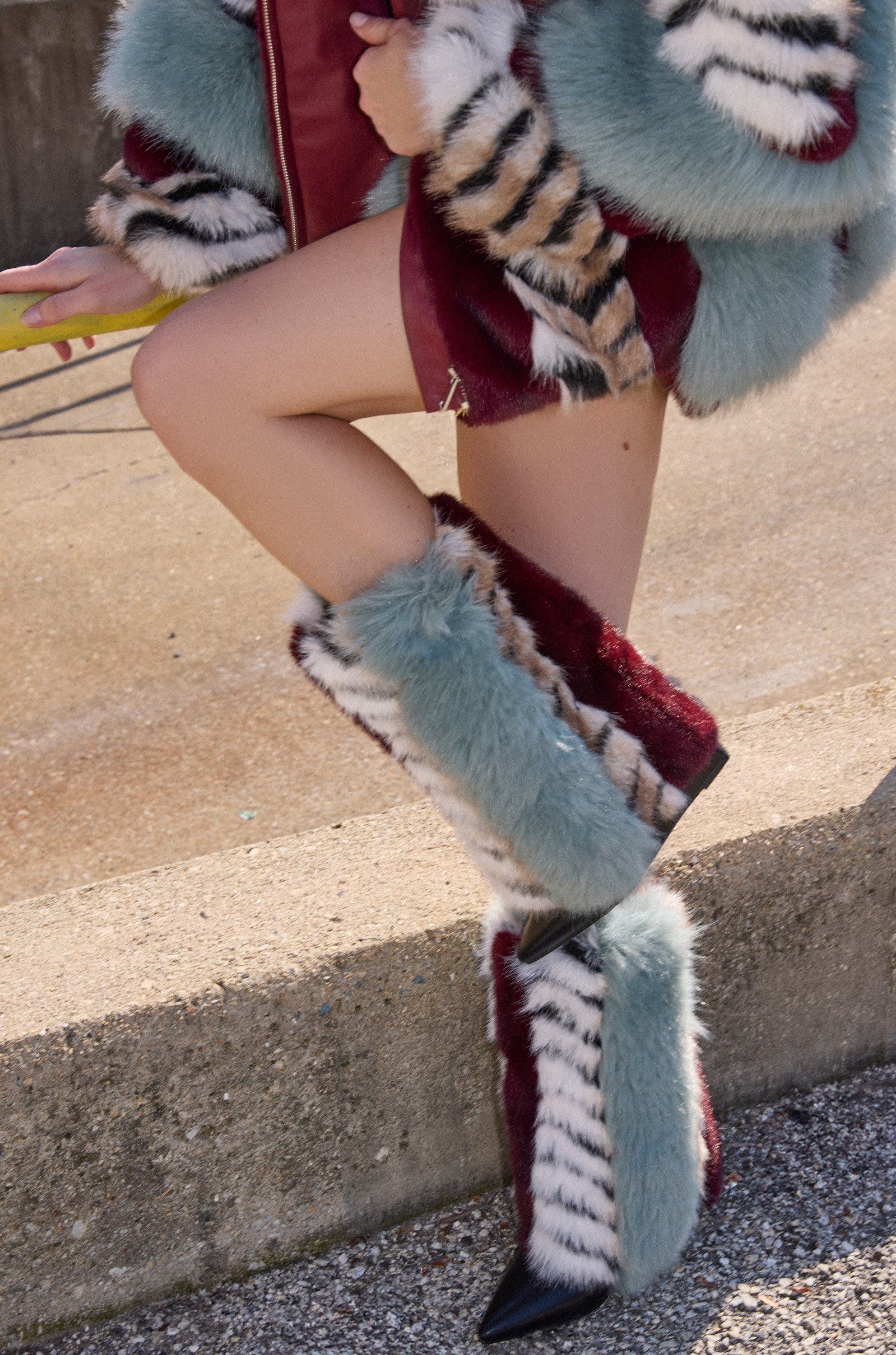A person stands outdoors on concrete, showing only their legs and outfit while wearing the AZALEA WANG BRIASH BLUE MULTI FUR HIDDEN WEDGE BOOTIE featuring blue, burgundy, white, and black tiger-like stripes.