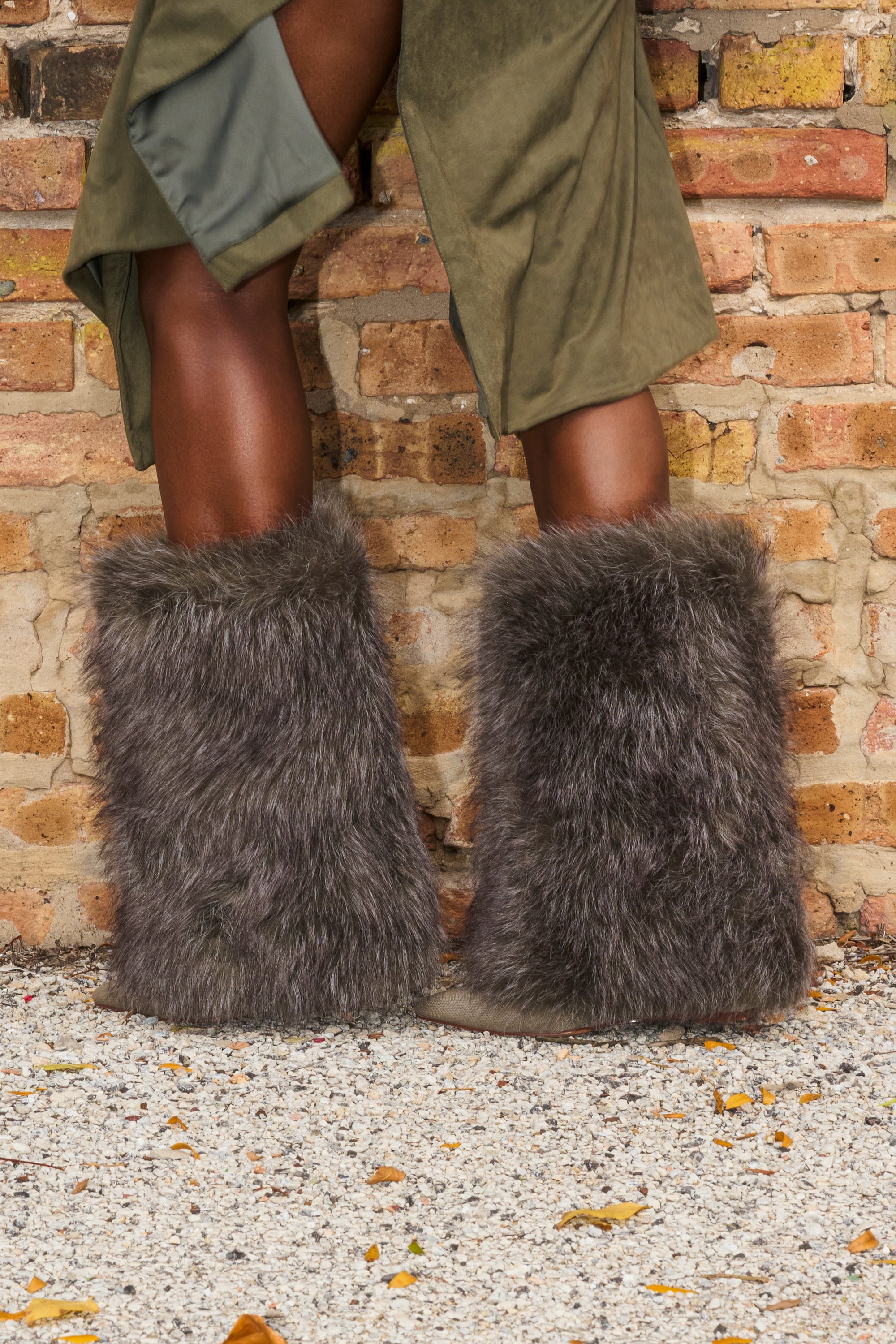 A person stands outdoors on gravel near a brick wall, wearing the AZALEA WANG BRIASH OLIVE WEDGE FUR BOOTIE with part of an olive green skirt or coat visible.