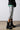 A person wearing a silver metallic knee-length skirt, AZALEA WANG BRIDGEY BLACK CHUNKY BOOTIE boots, and a black and green varsity jacket is captured standing on a neutral floor against a plain background.