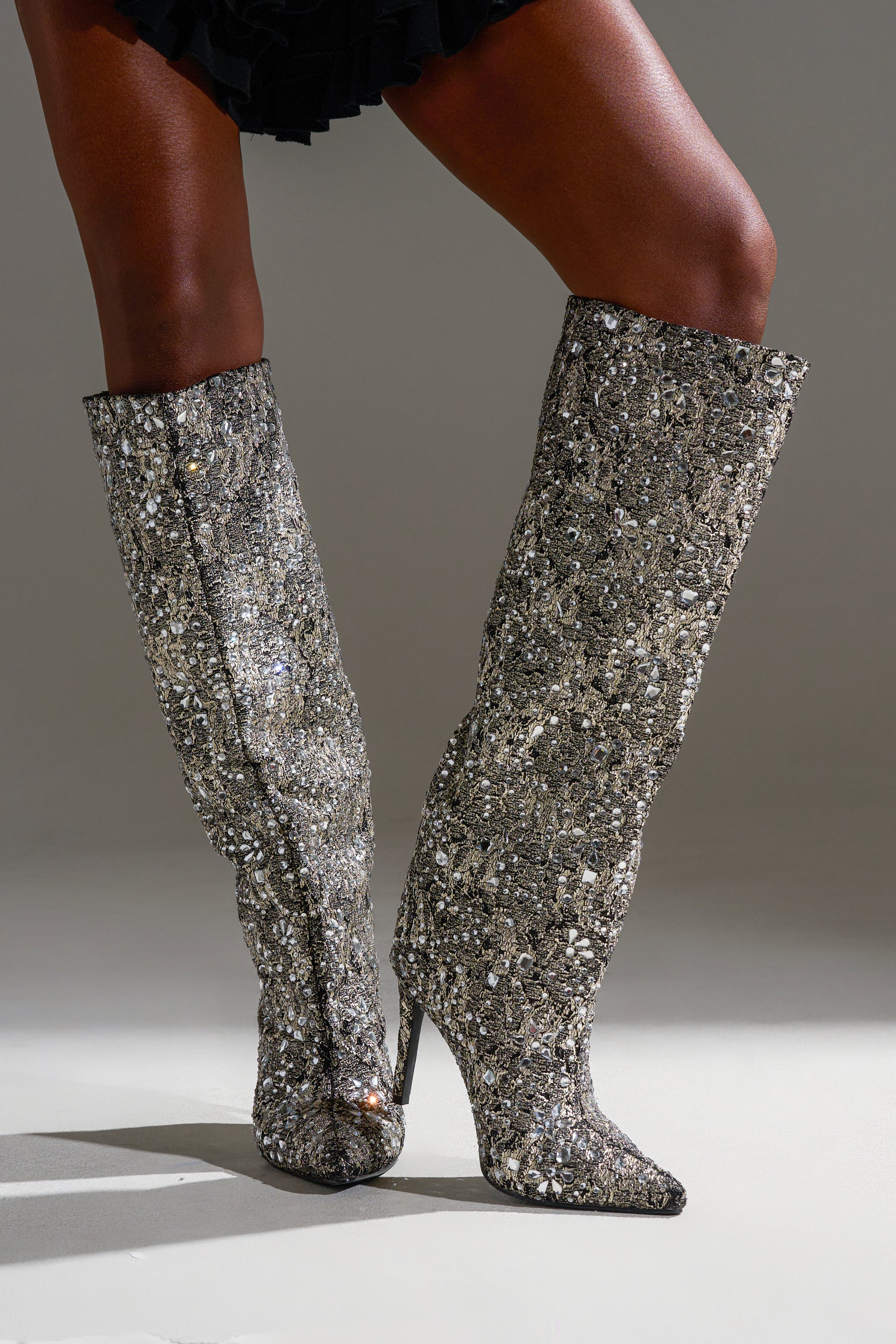 A person stands with legs crossed in AZALEA WANG BRIELEY GOLD AND BLACK FLORAL BROCADE KNEE HIGH BOOTS, their ornate pointed toes and stiletto heels shining under the light, paired stylishly with a short black skirt.