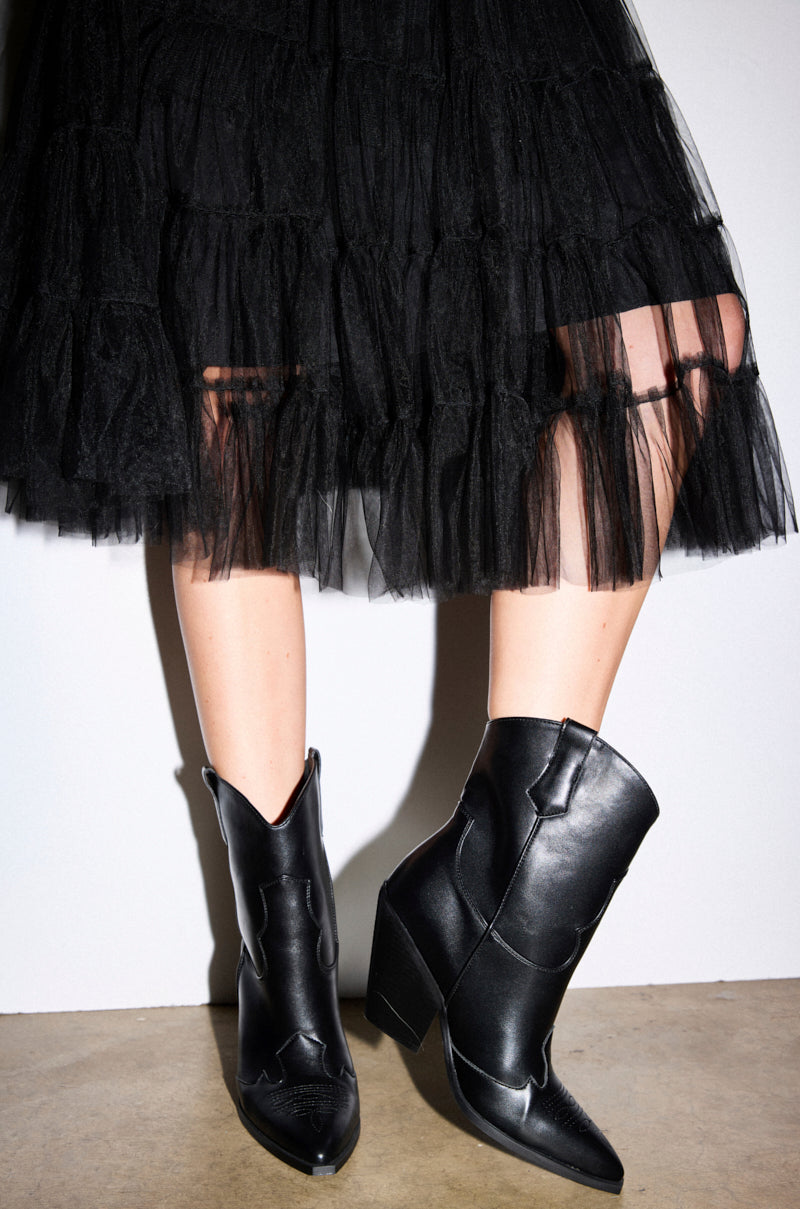 Someone stands on a concrete floor against a white wall, wearing a black tulle skirt and AZALEA WANG BRIGGITA BLACK WESTERN COWBOY BOOTIE. Only their lower legs and feet are visible. 2