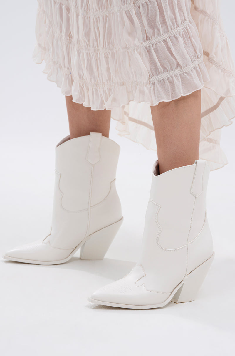 A person in a light, ruffled skirt wearing AZALEA WANG BRIGGITTA WHITE WESTERN COWBOY BOOTIE with pointed toes and angled heels stands on a white surface.