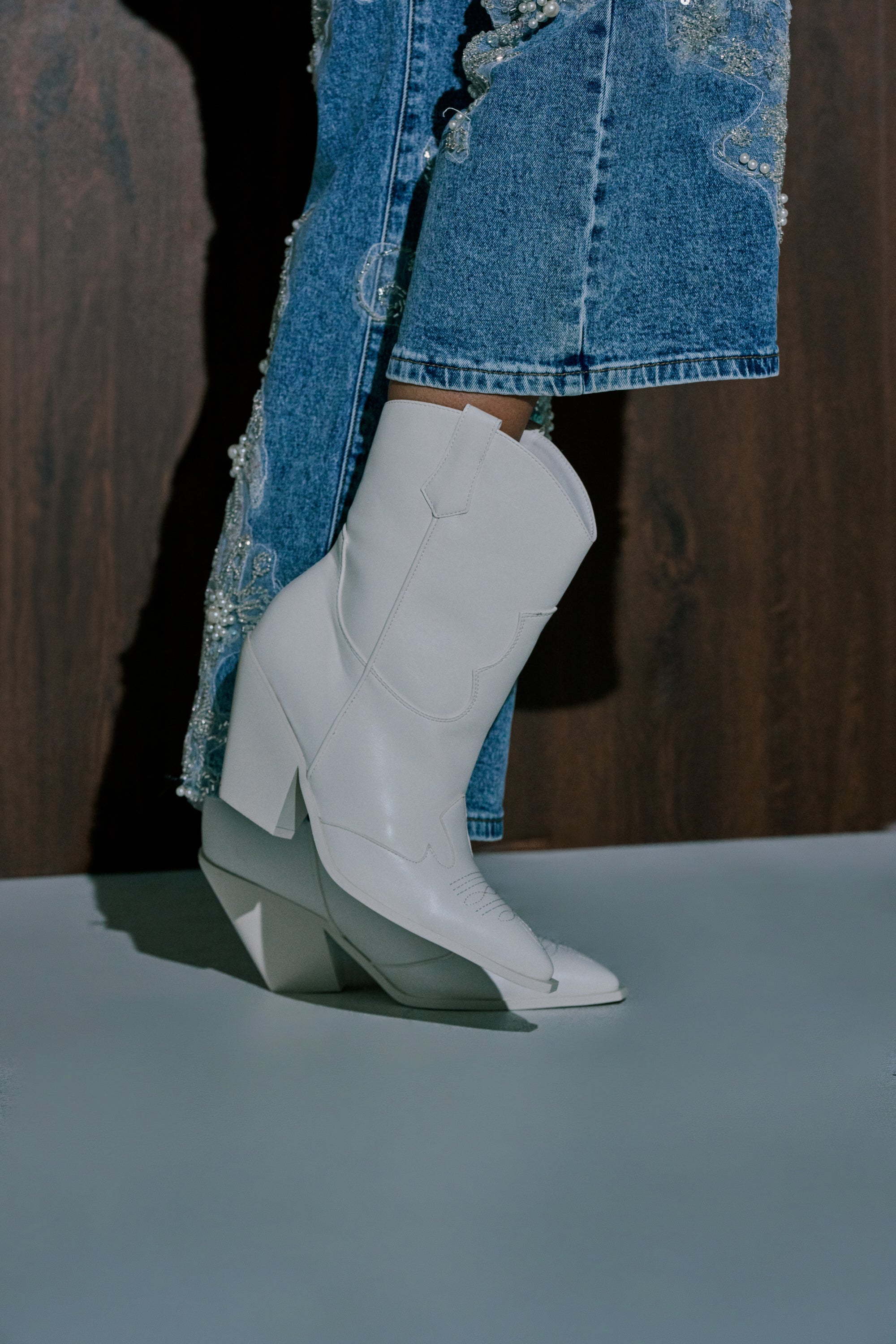 A person in embroidered blue jeans and AZALEA WANG BRIGGITTA WHITE WESTERN COWBOY BOOTIES stands against a dark wooden background. 0