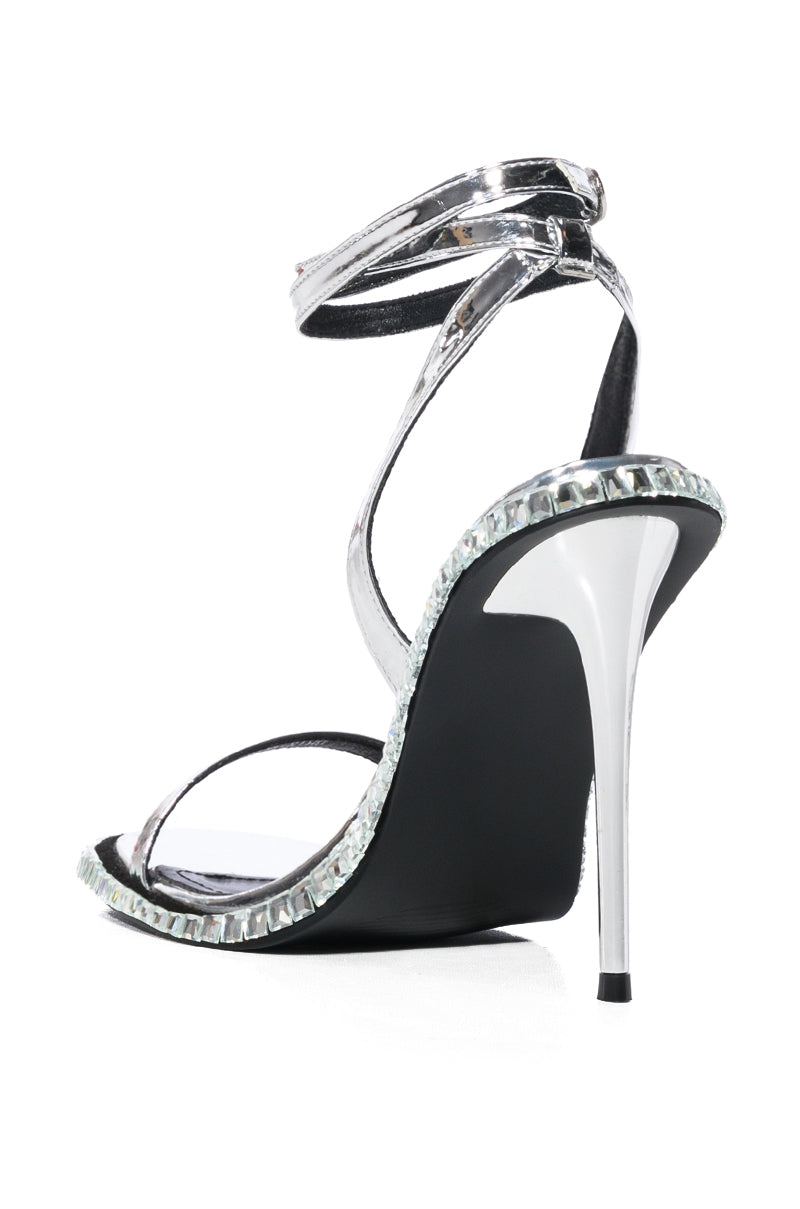 The AZALEA WANG BRIK BLACK stiletto sandal showcases a shiny silver strap with an ankle buckle. The back view highlights the faux leather heel and edge, adorned with rhinestone trim for a sparkling, elegant design.