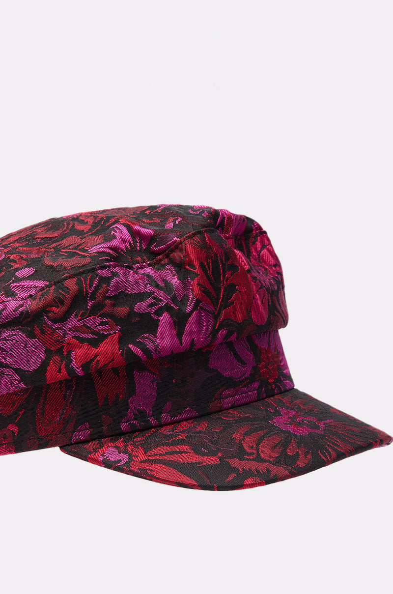 A close-up of the LUXE LIVING BRIM HAT reveals its vibrant floral brocade pattern in red, purple, and black, set against a plain light background, highlighting its stylish flat brim design.