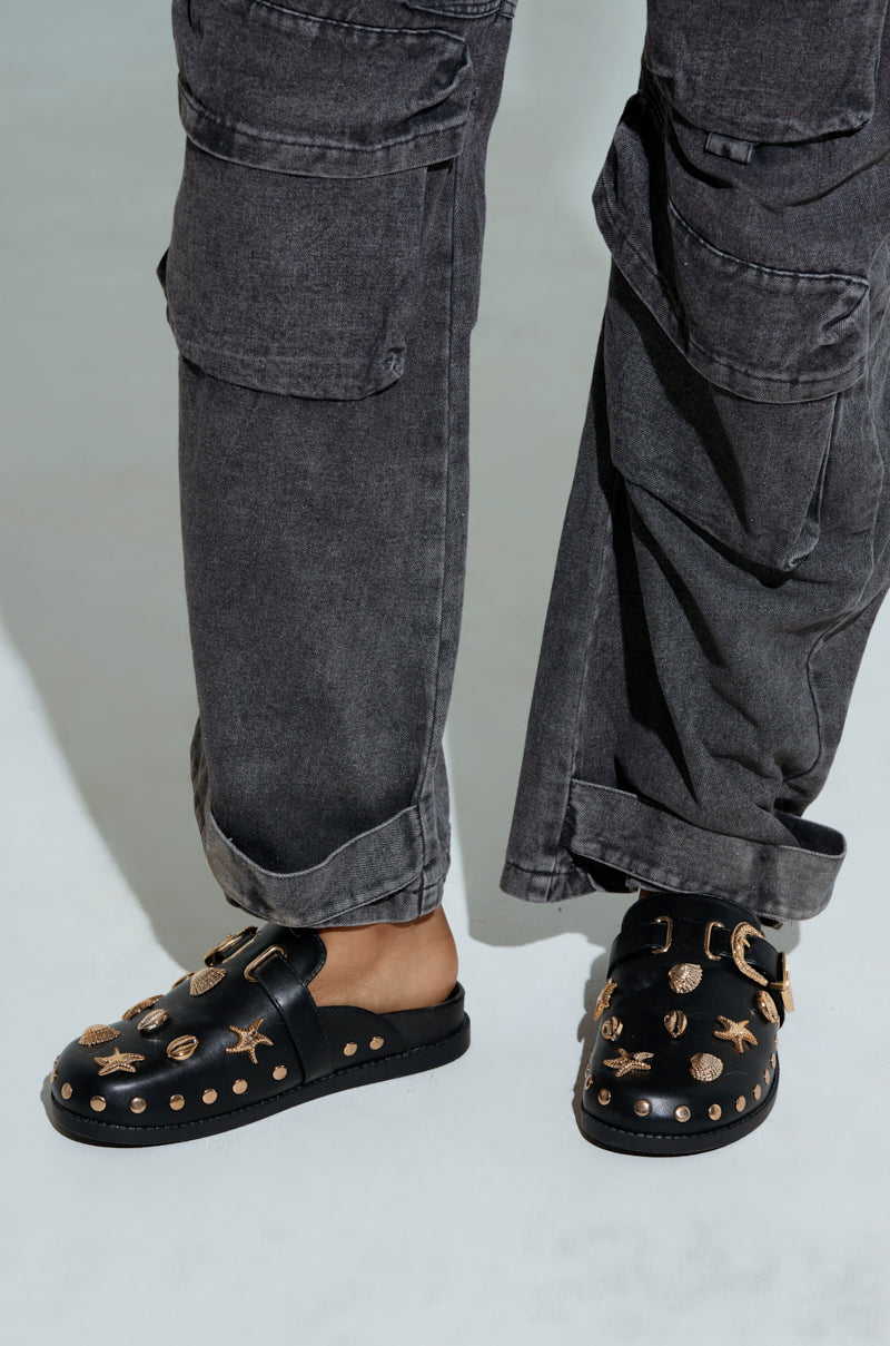 A person wears black pants with a relaxed fit and multiple pockets, paired with the AZALEA WANG BRINY BLACK SEA SHELL EMBELLISHED CLOG, showcasing chic black slip-on shoes adorned with gold and sea shell accents. The look is set against a smooth, light background.