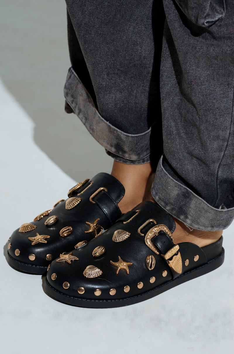 A person wears the AZALEA WANG BRINY BLACK SEA SHELL EMBELLISHED CLOG, featuring black vegan leather with almond toes and adorned with gold seashells and starfish. Gold studs and buckles enhance the design, while rolled-up jeans perfectly highlight these artistic details.