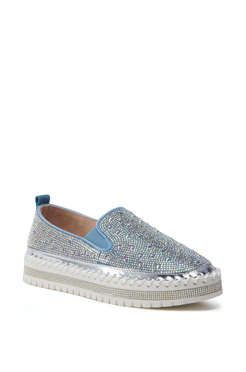 A single AZALEA WANG BRITTANY DENIM RHINESTONE SNEAKER is displayed against a plain white background. The sneaker features numerous small, glittering rhinestones and boasts a thick white flatform sole with silver detailing around the edge. This shoe has a casual yet glamorous design.