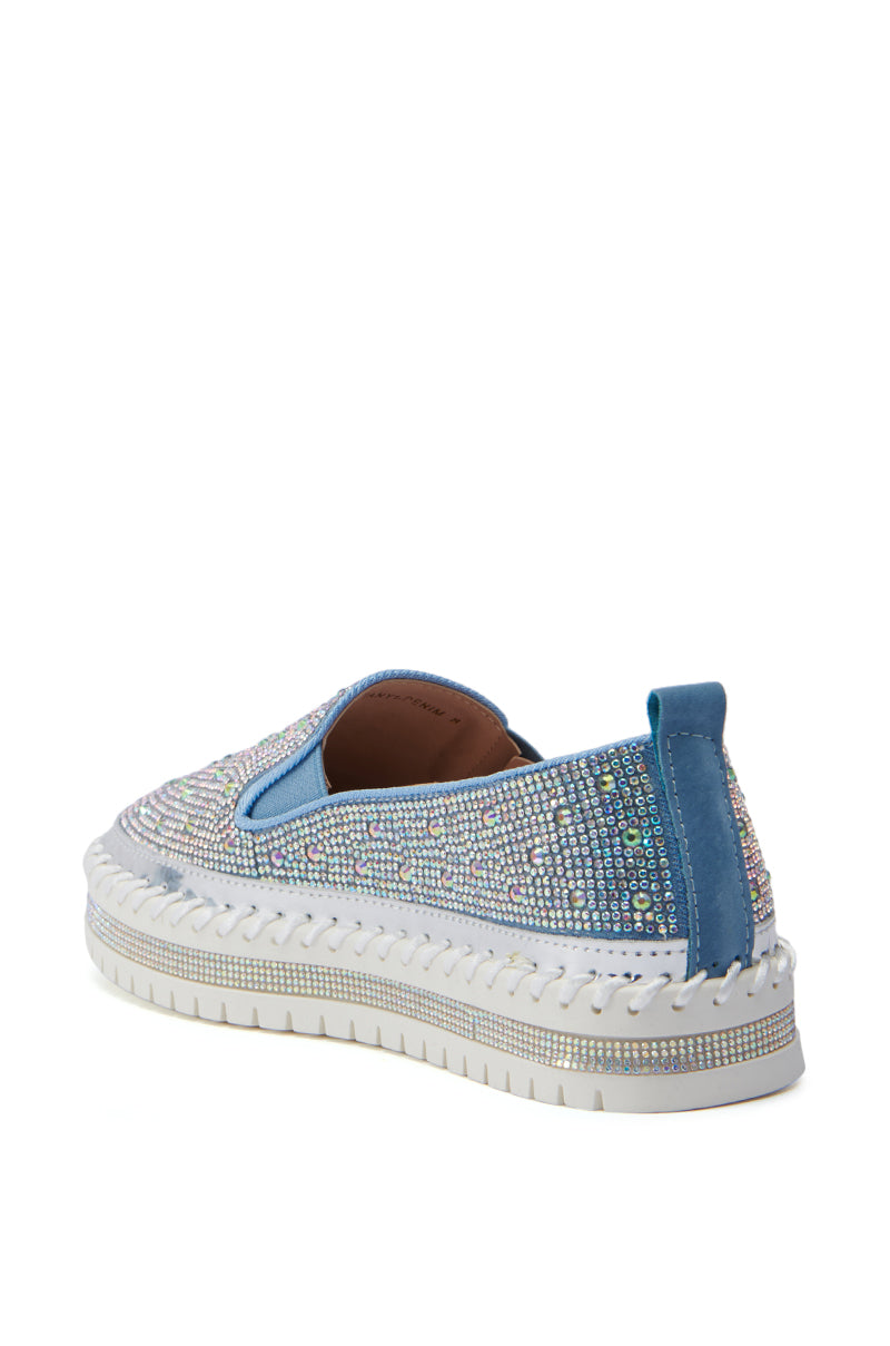 The AZALEA WANG BRITTANY DENIM RHINESTONE SNEAKER features a single blue slip-on design adorned with numerous shiny rhinestones. It boasts a faux leather upper, a thick flatform sole with intricate stitching details, and a pull tab at the back for easy wear. The angle is from the back-left side, highlighting the detailed decoration.