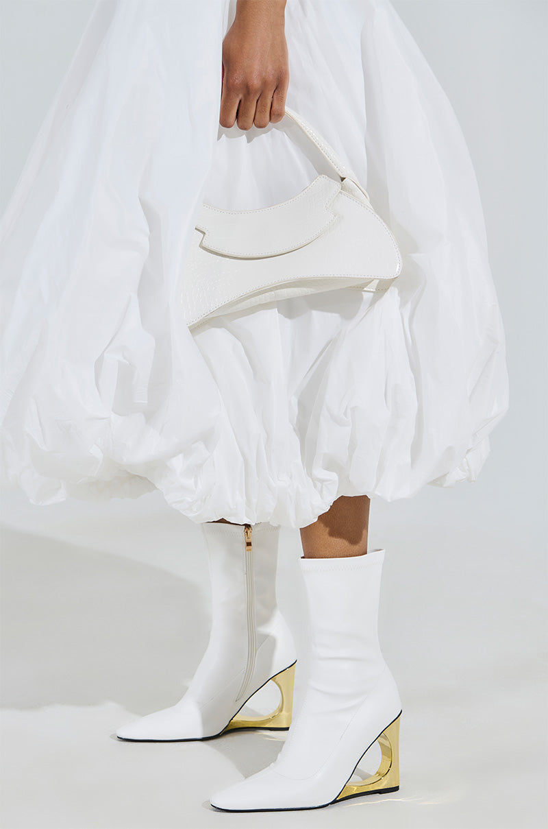 A person in a voluminous white dress and AZALEA WANG BRIXHAM WHITE BOOTIE WITH NOVELTY HEEL, featuring pointed toes and gold accents, holds a white handbag. The minimalist setting with a light backdrop heightens the ensemble's elegance.