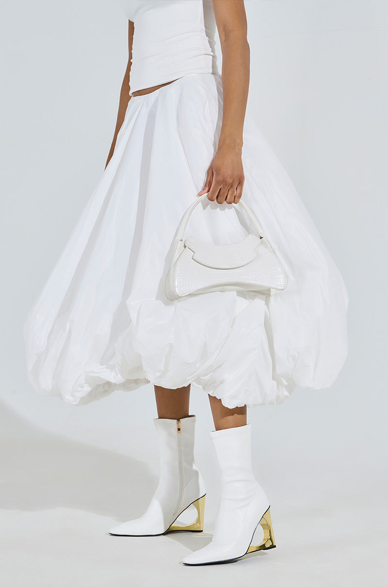 Dressed in white, a person models a voluminous skirt and top against a plain backdrop. They accessorize with the AZALEA WANG BRIXHAM white booties featuring gold accents and a novelty heel, while holding a small white handbag.