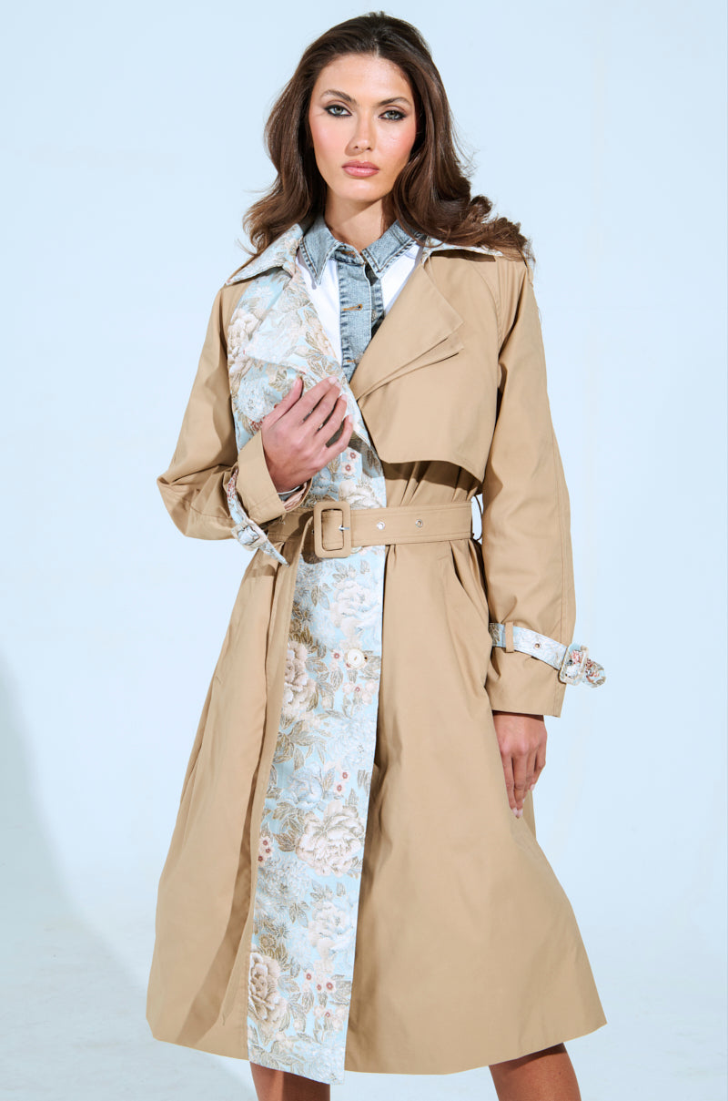 Wearing the PEEKABOO BROCADE TRIM TRENCH, a person stands against a light blue background. The beige trench coat features vintage floral and denim details, with brocade trim accentuating the belted waist. Their long brown hair and calm expression complete the look.