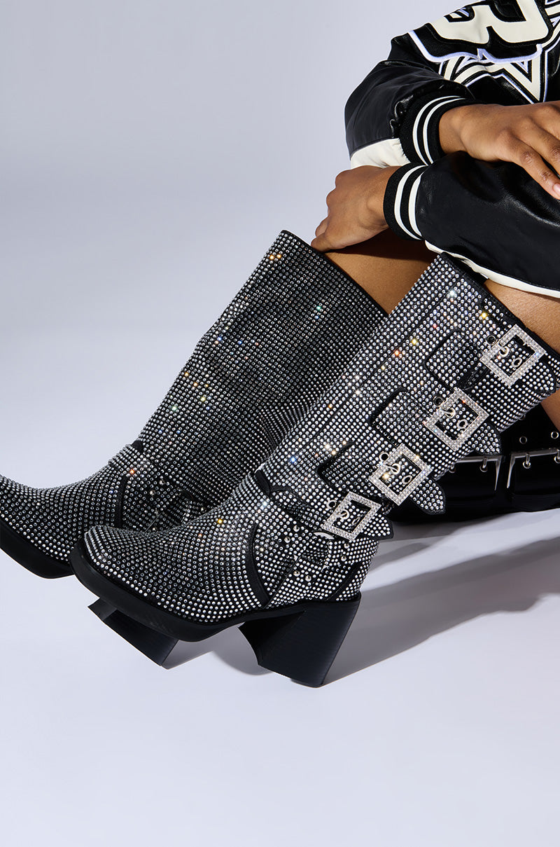A person wearing the AZALEA WANG BRONSON BLACK RHINESTONE EMBELLISHED BOOT—knee-high, high-heeled black boots adorned with sparkling rhinestones and featuring multiple decorative silver buckles along with harness-inspired embellishments. They are paired with a black and white varsity-style jacket as the person poses against a plain background. 1