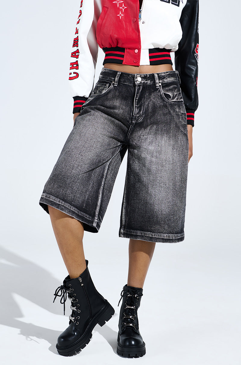A person is dressed in a red and black varsity jacket, gray knee-length denim shorts, and the sleek AZALEA WANG BROOKS BLACK COMBAT BOOTIE against a plain light gray background.