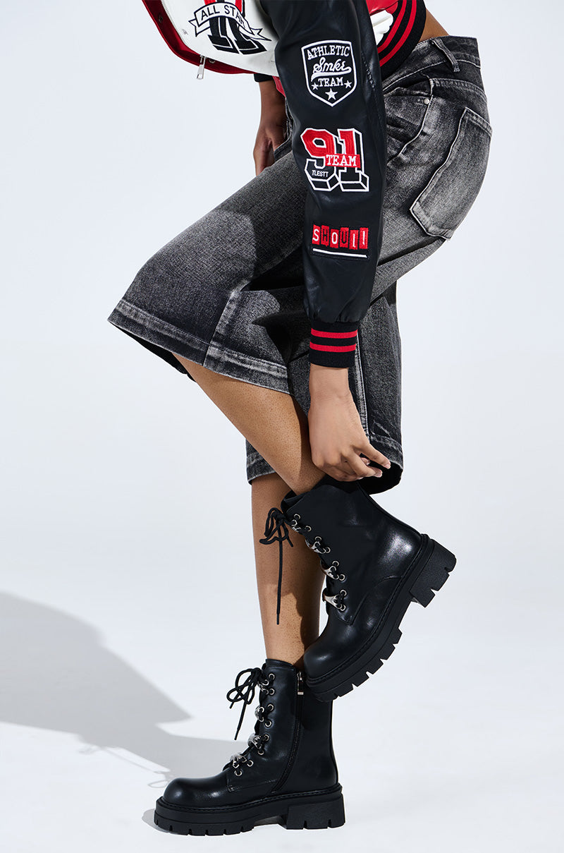 A person adjusts the lace of an AZALEA WANG BROOKS Black Combat Bootie while wearing dark denim shorts and a black jacket with red and white patches against a pure white backdrop.