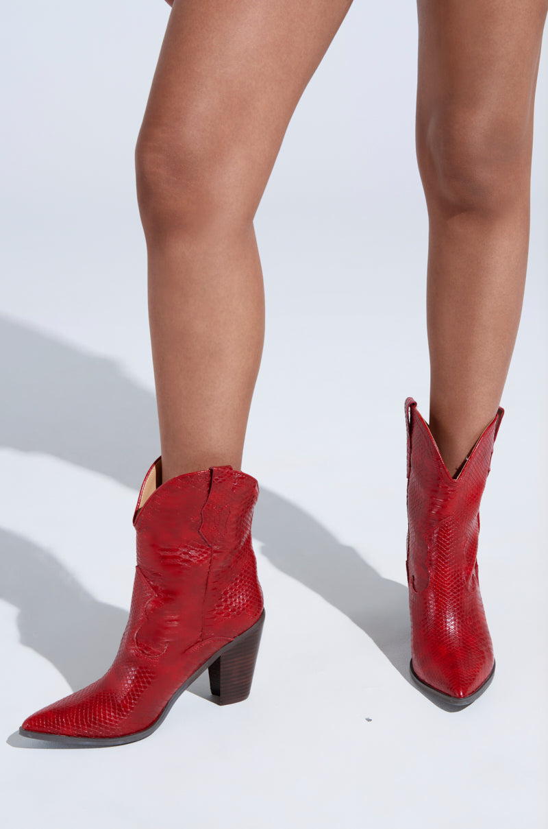 A person wearing the Brooklyn Red Western Cowboy Bootie with a textured design and chunky heels stands on a light gray floor, spotlighting the bold color and pointed-toe style of the footwear. 0