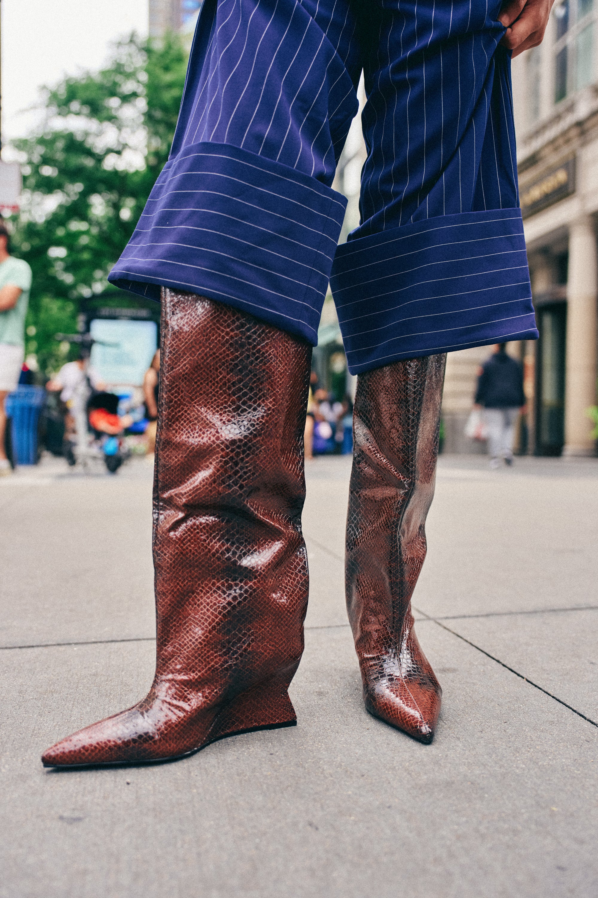 Wearing blue pinstripe pants, a person stands on a city sidewalk in the AZALEA WANG BRUN BROWN SNAKE WEDGE BOOT—a knee-high brown snakeskin-print boot with a slim wedge heel.
