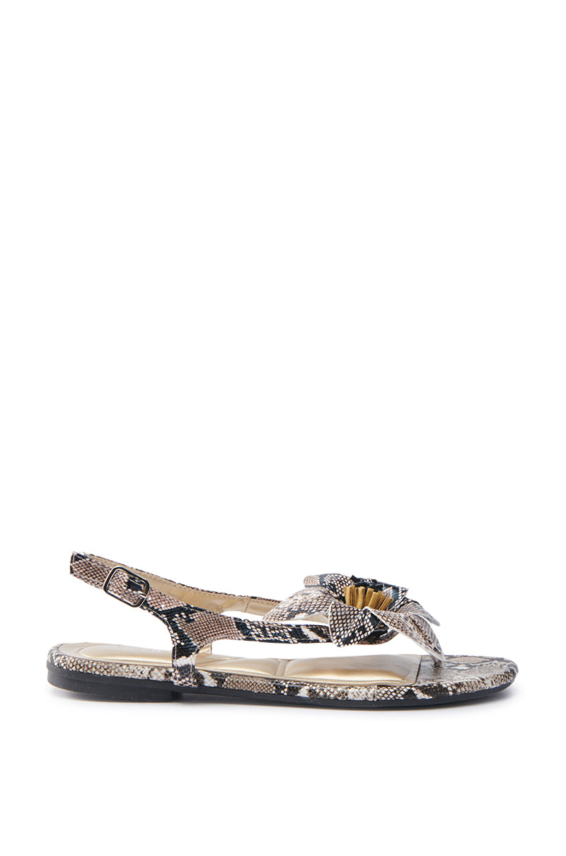 The AKIRA BRUNA SNAKE SANDAL is a chic sandal with a glossy faux snakeskin pattern, flat sole, and slingback strap. It features a front knot detail and an adjustable buckled ankle strap against a plain white backdrop to enhance its sleek design.