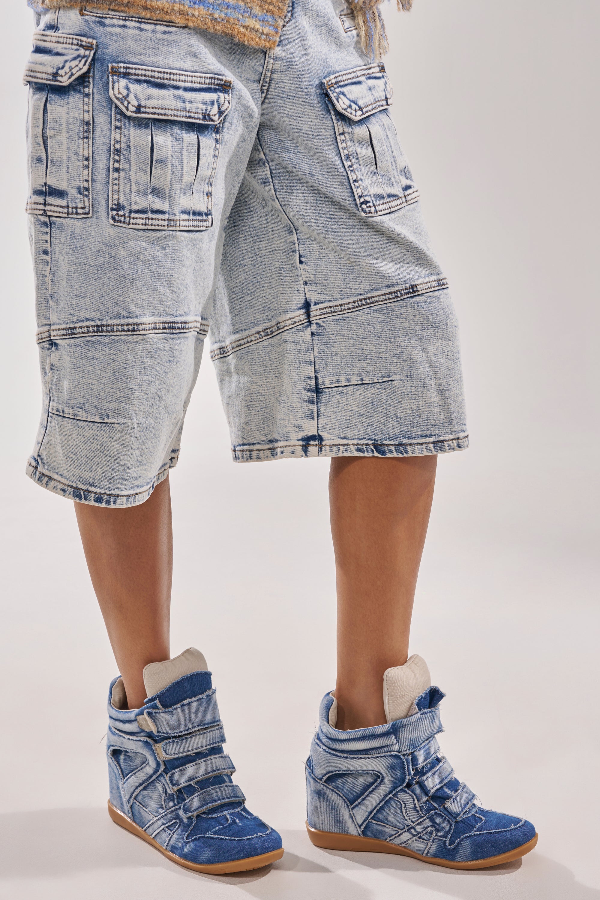 Wearing knee-length, light-washed denim cargo shorts and AZALEA WANG BRYANT DENIM WEDGE SNEAKER with velcro straps, a person stands against a plain light background. 0
