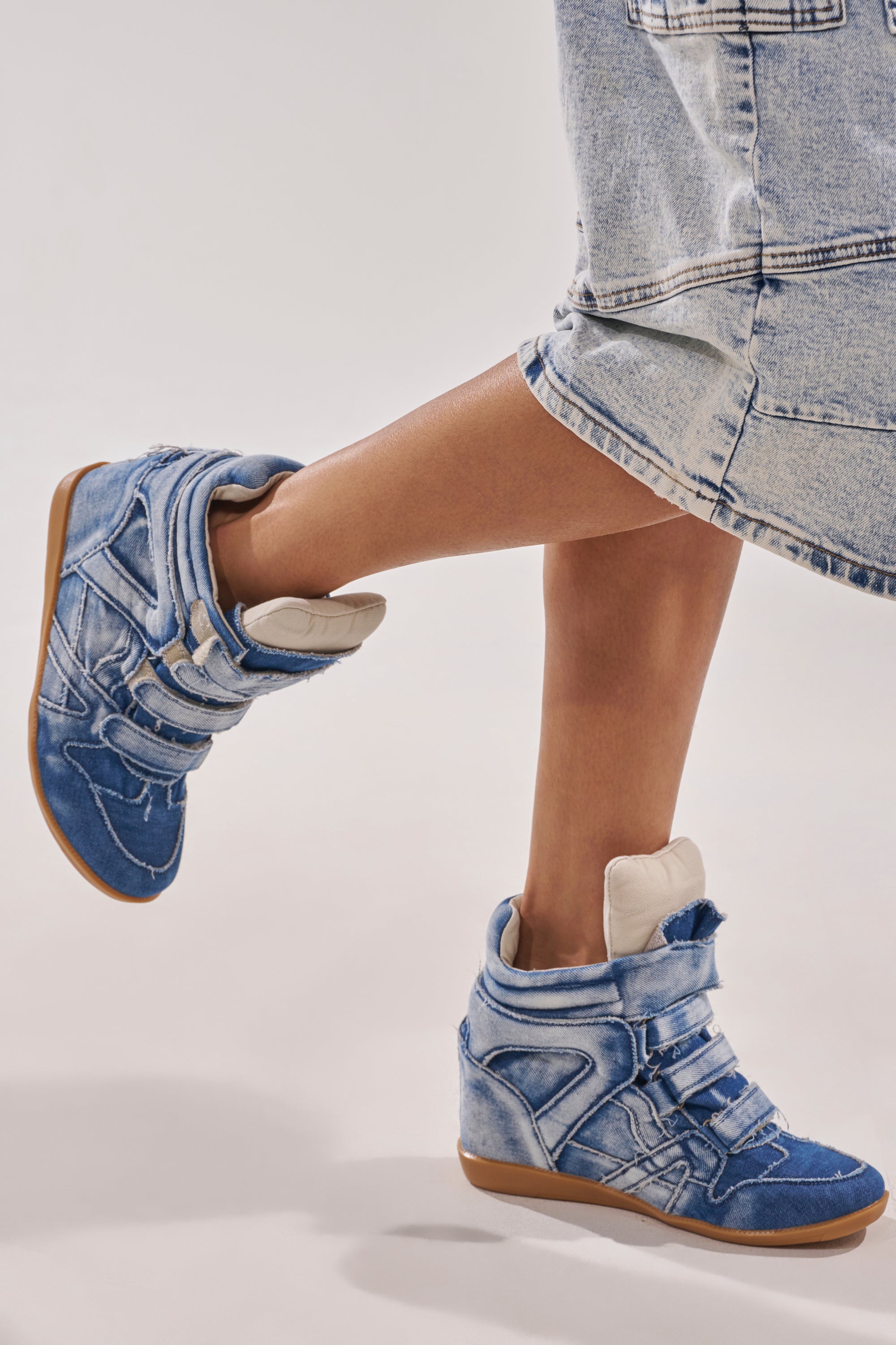 A person wearing a light denim skirt and AZALEA WANG BRYANT DENIM WEDGE SNEAKERS with velcro straps and gum soles, standing on tiptoe against a plain background. 1