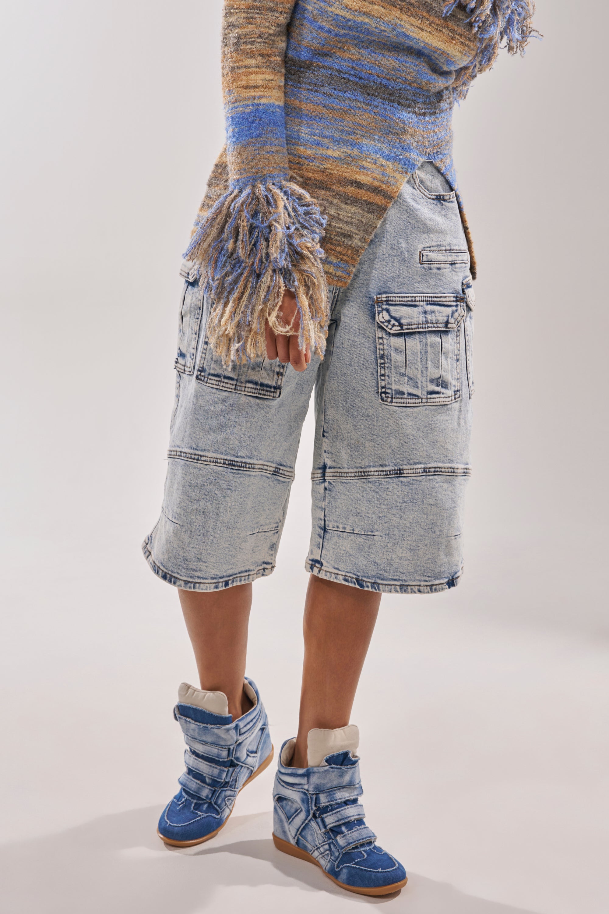 Wearing the AZALEA WANG BRYANT DENIM WEDGE SNEAKER, multicolored fringed sweater, and loose light-wash denim shorts with large pockets—a trendy streetwear look cropped from shoulders down. 7